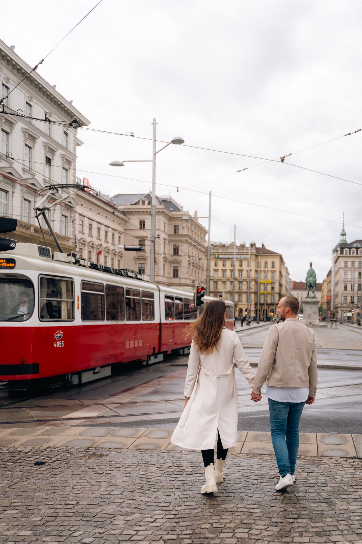 Couple holding hands walking near iconic red Vienna tram on cobblestone city street.