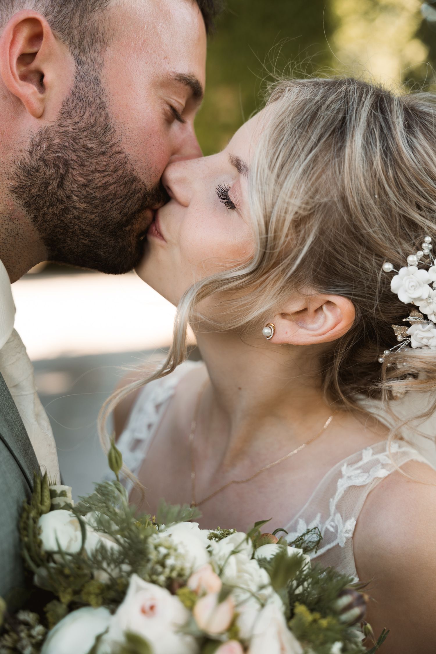 Close-up of bearded groom kissing bride holding a lush white and green floral bouquet.