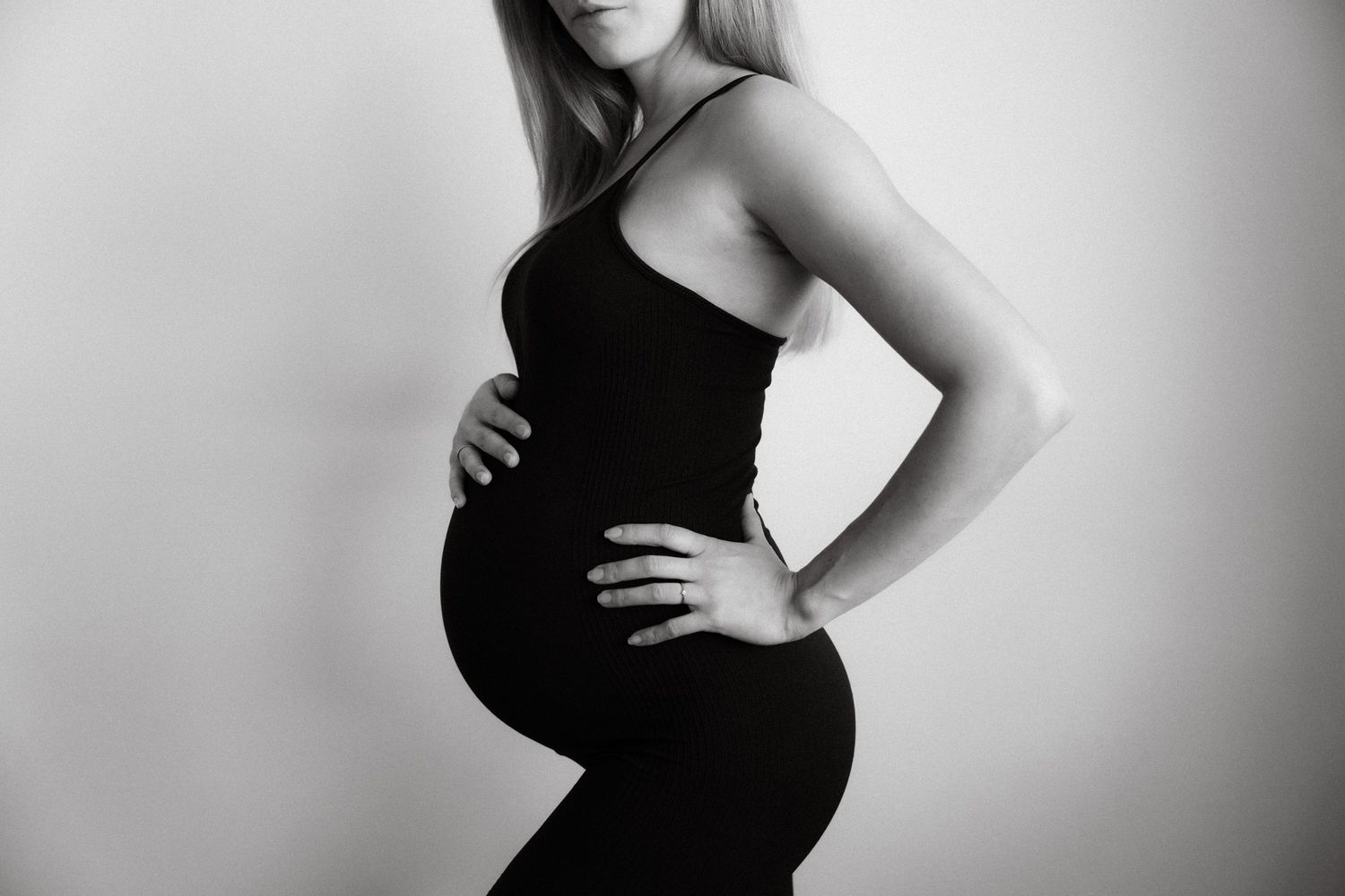 Black and white profile photo of pregnant woman in black dress, hands on hips, showcasing baby bump.