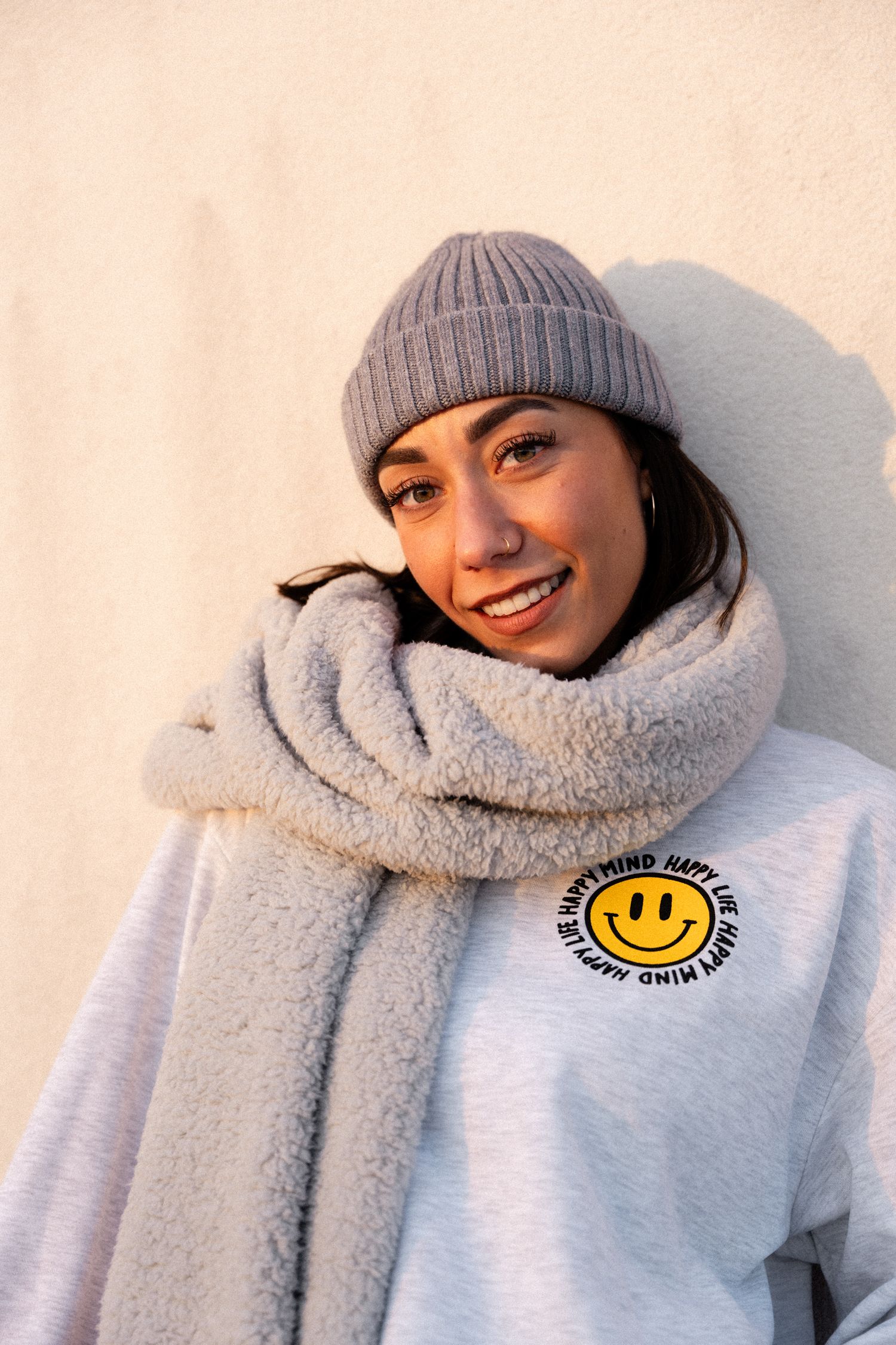 Smiling young woman wearing a gray beanie hat, fluffy scarf, and smiley face sweatshirt leaning against a wall.