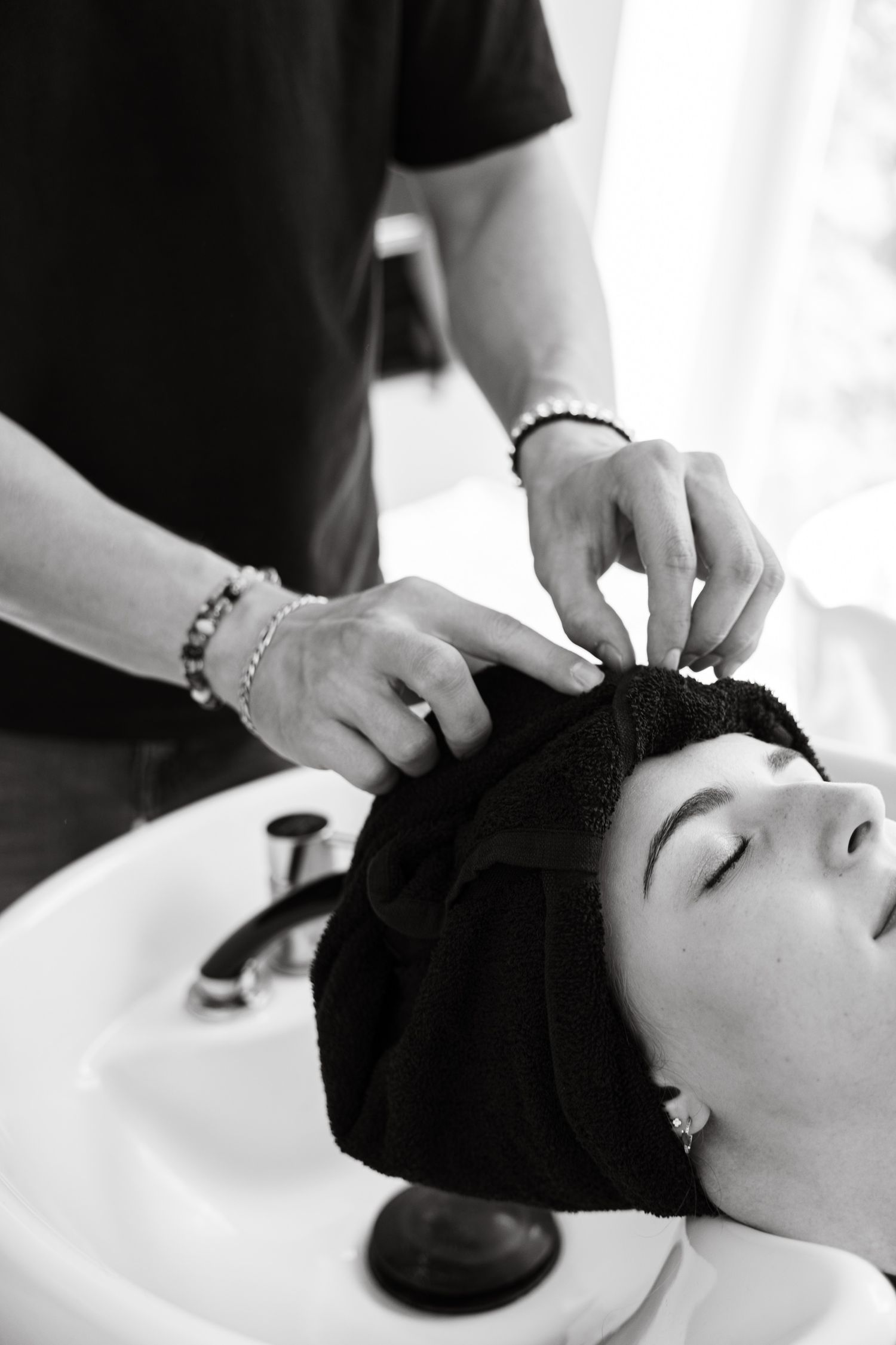 Hair stylist wrapping client's wet hair in towel at salon shampoo bowl.