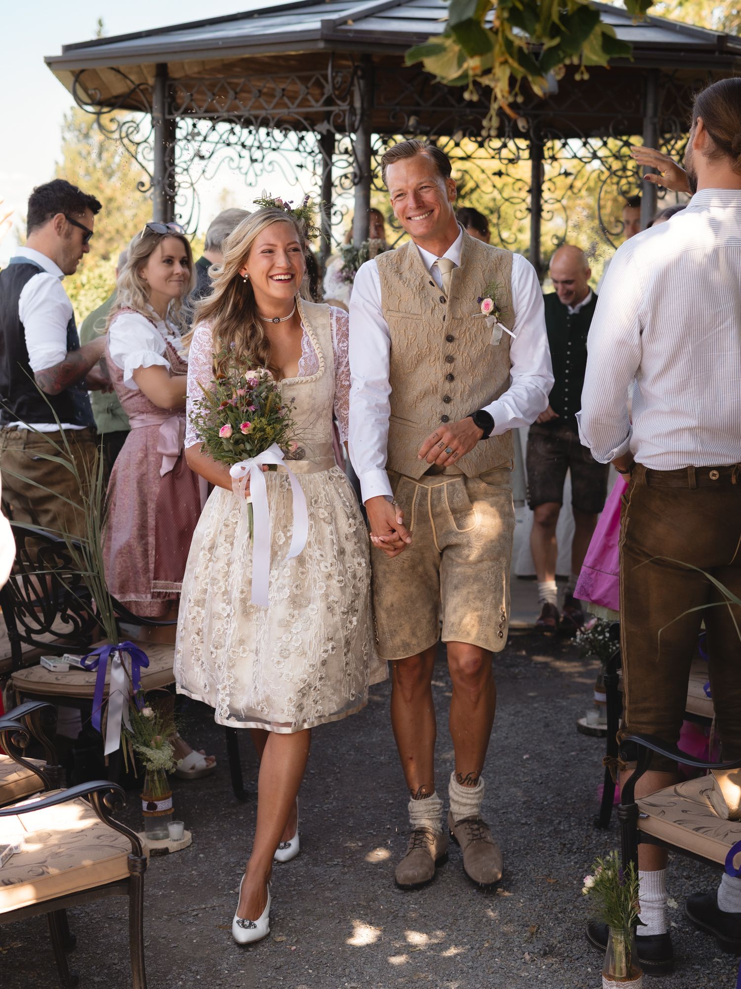Couple in traditional Bavarian dirndl and lederhosen walking joyfully through outdoor wedding aisle.