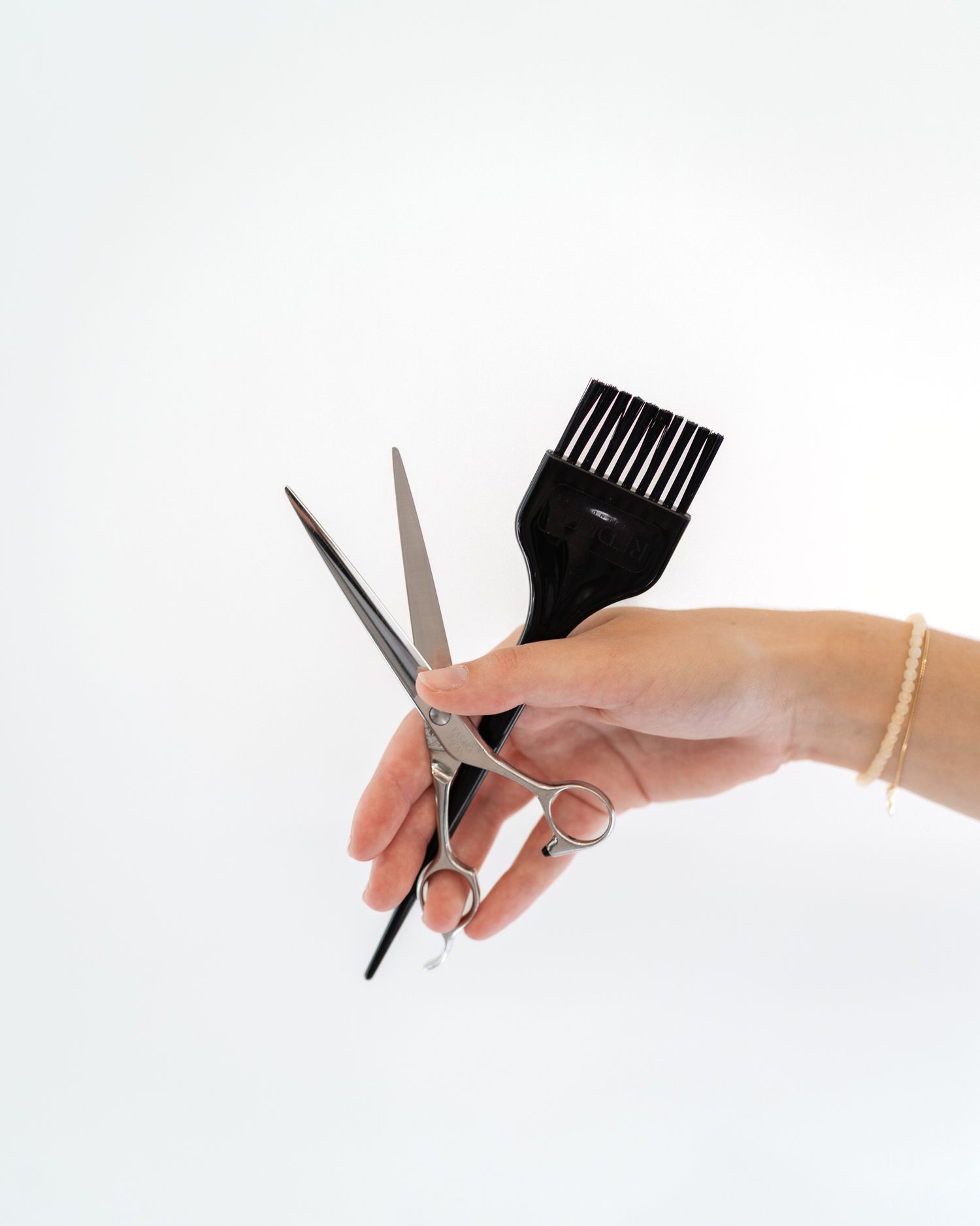 Hand holding professional hair cutting scissors and black dye brush against white background.
