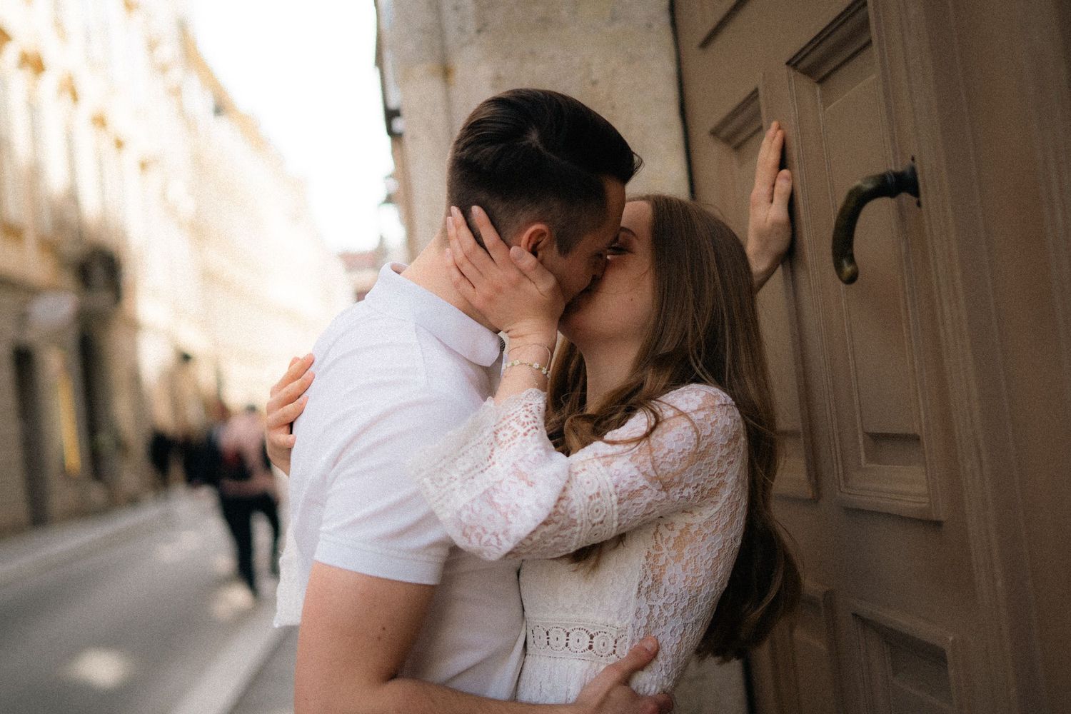 Couple sharing a romantic kiss beside a wooden door on a sunlit European cobblestone street.