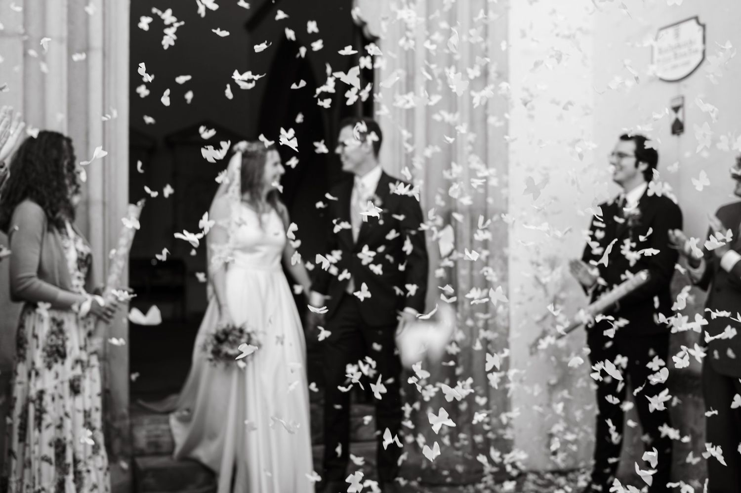 Black and white photo of newlyweds exiting church surrounded by white butterfly confetti with guests celebrating.