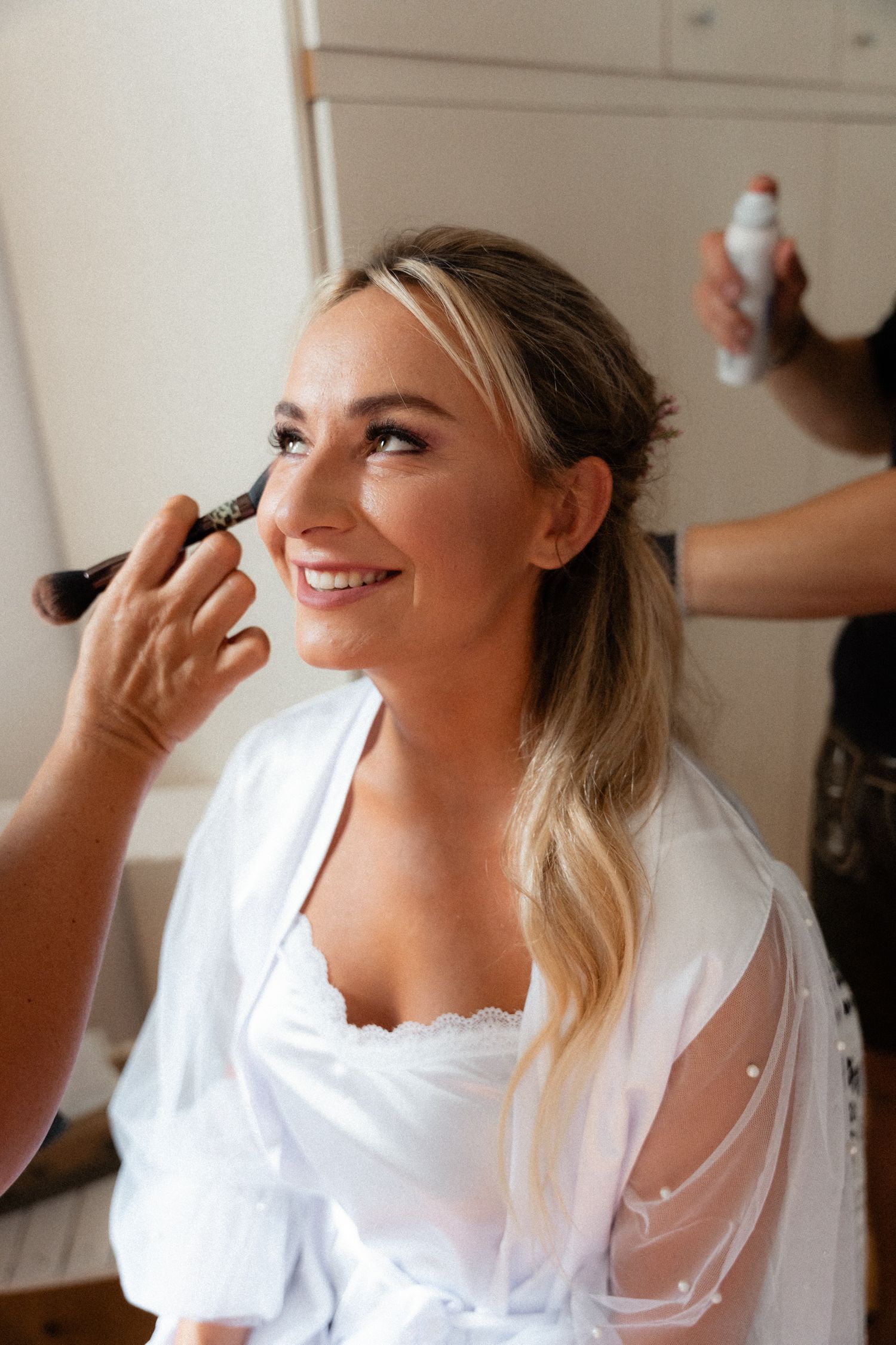 Smiling blonde bride in white robe getting makeup applied by professional artist before wedding.