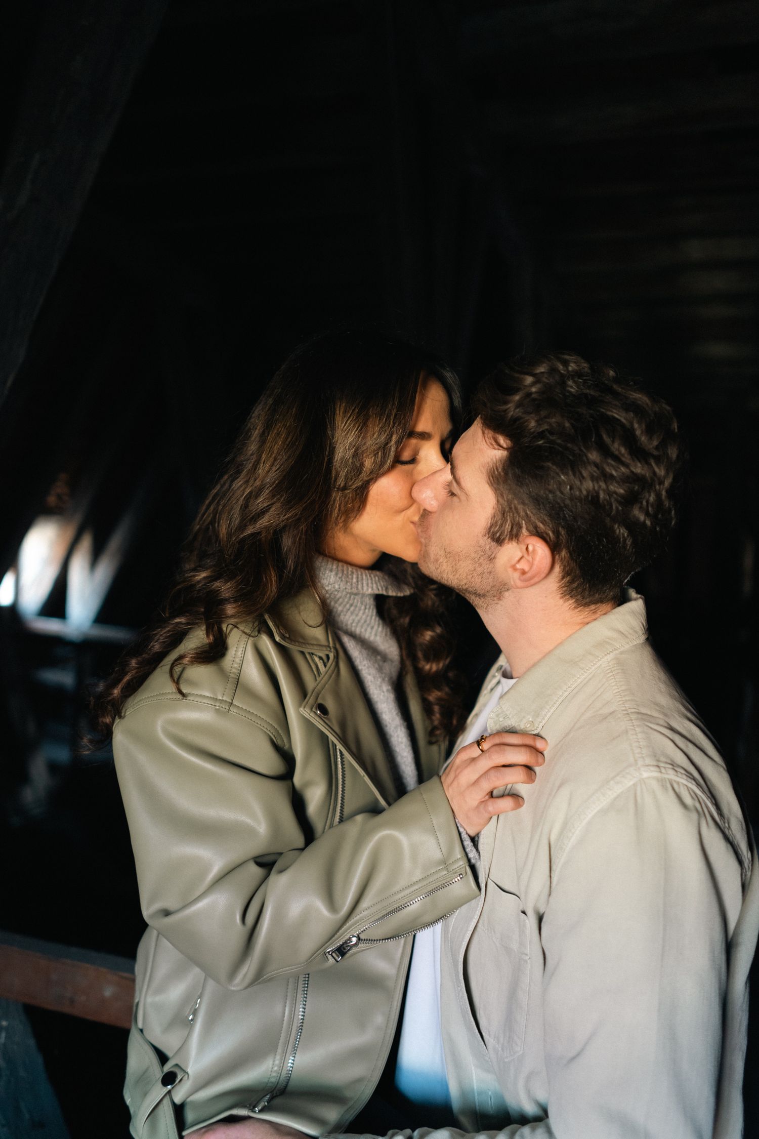Couple sharing a tender kiss in a dimly lit rustic setting, both wearing stylish leather jackets.