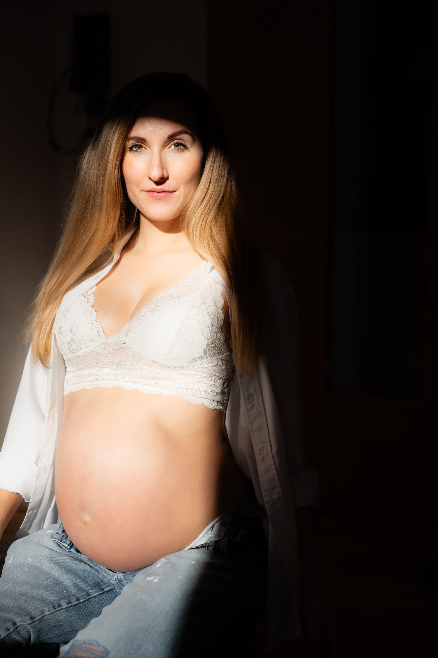 Pregnant woman in white lace bra and jeans wearing black hat posing in dramatic sunlight.