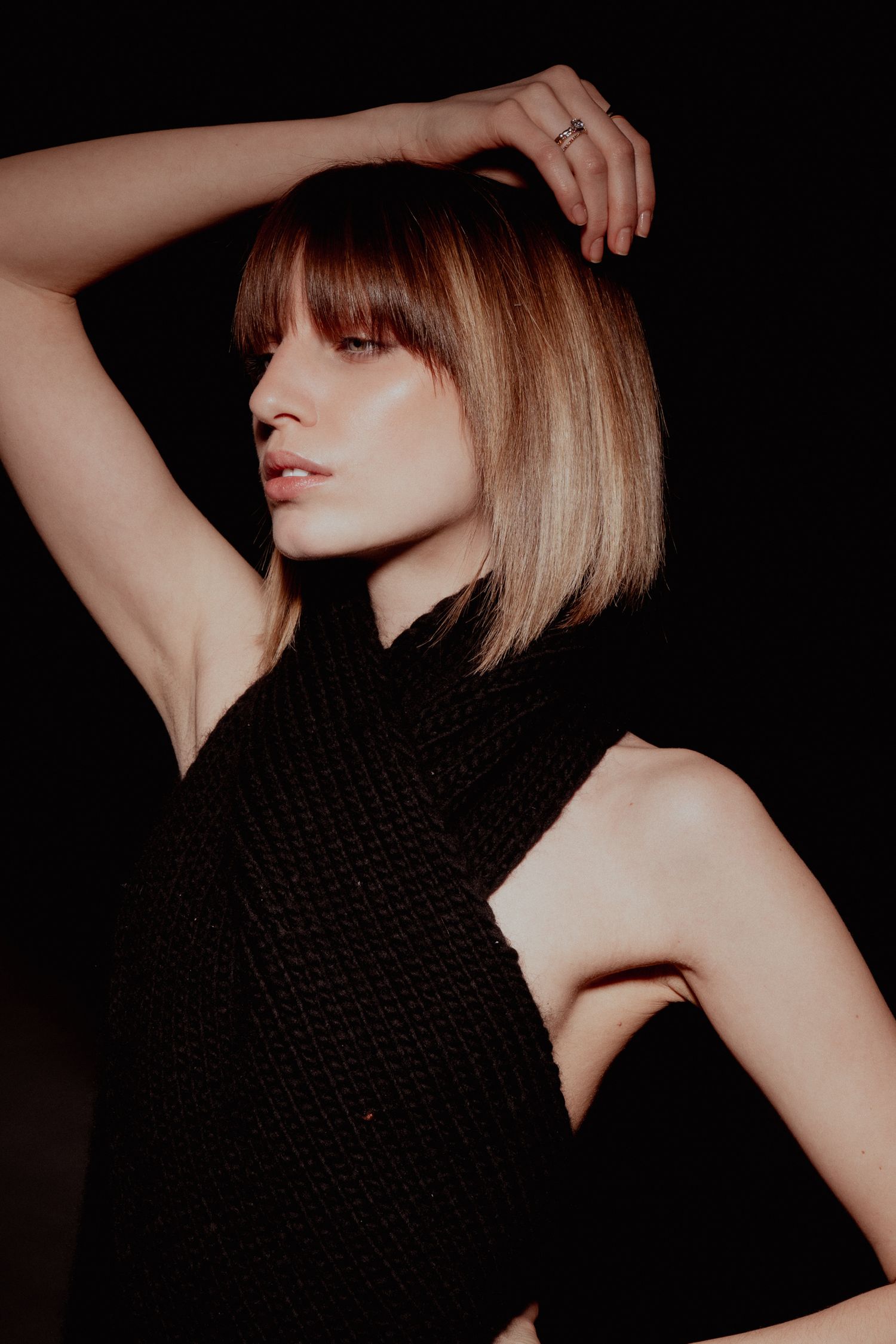 Artistic dark portrait of a woman with highlighted bob haircut in black sleeveless top raising arm overhead.