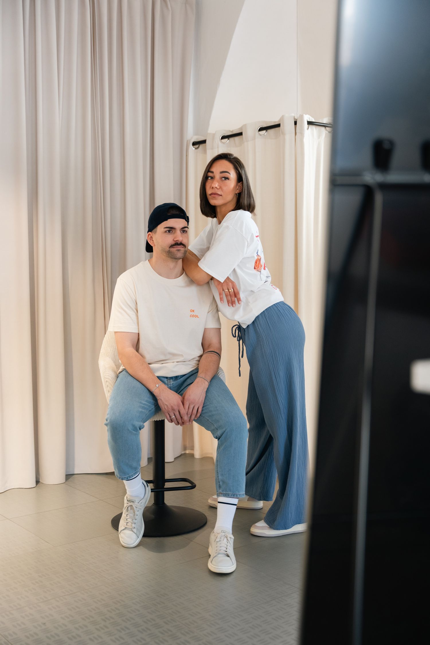 A man sitting on a stool and a woman standing beside him, both wearing casual white tops and blue jeans indoors.