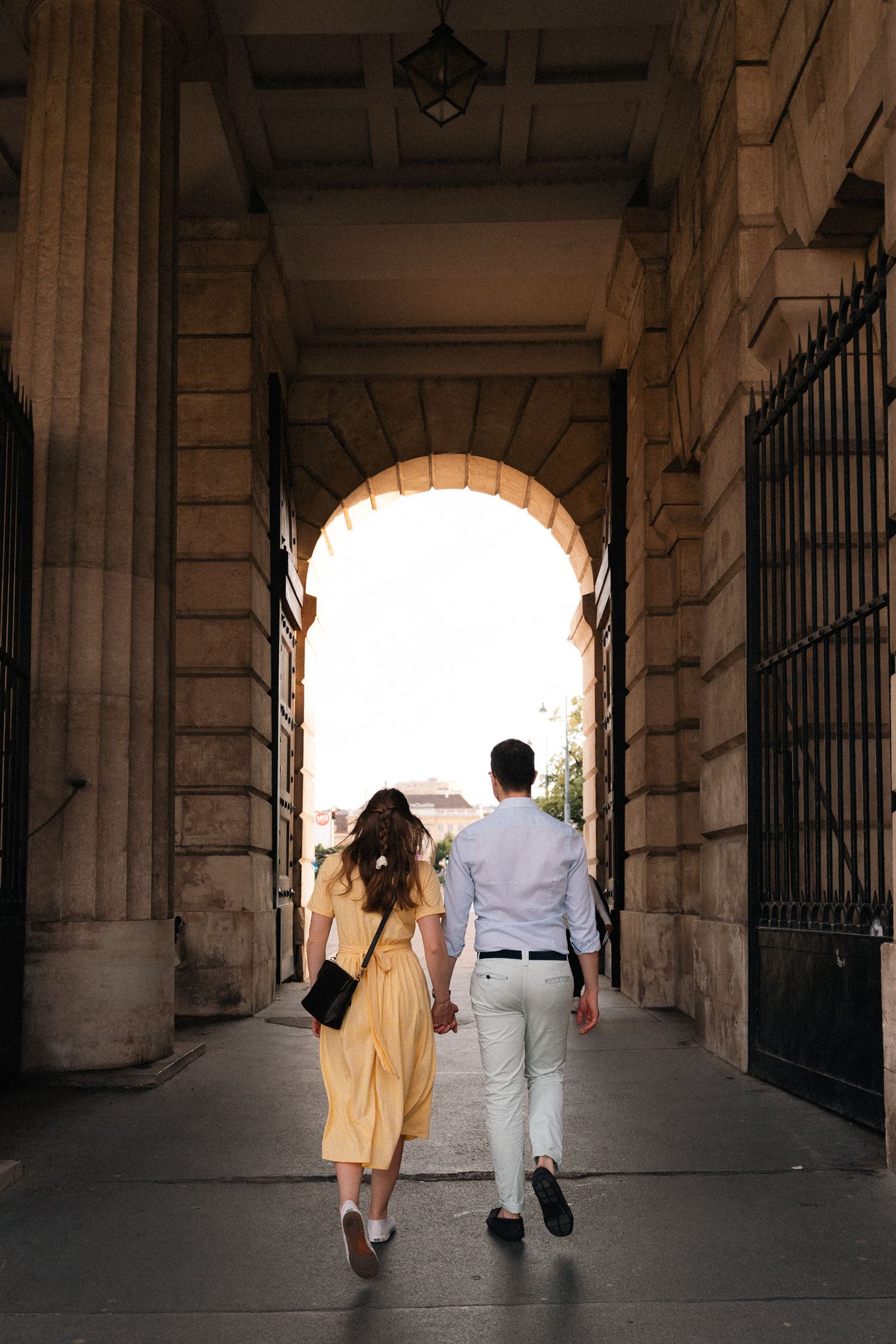 Couple holding hands walking through stone archway passage in historic European building.