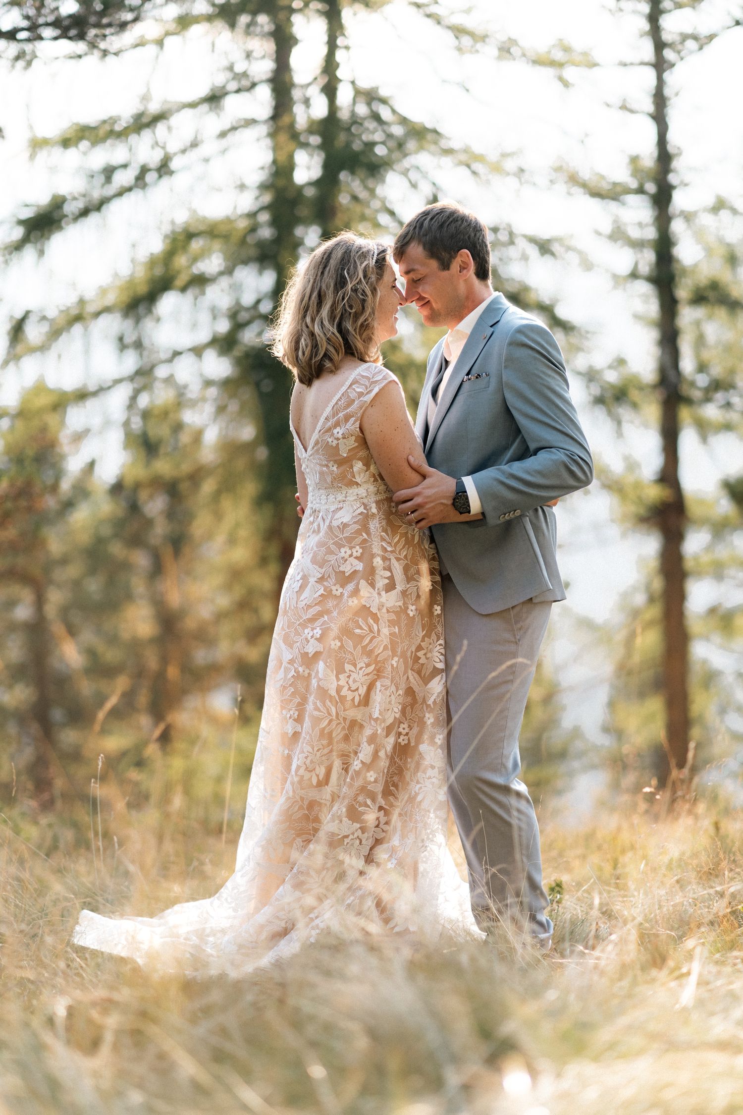 Bride in lace gown and groom in gray suit sharing a tender moment in golden forest meadow.