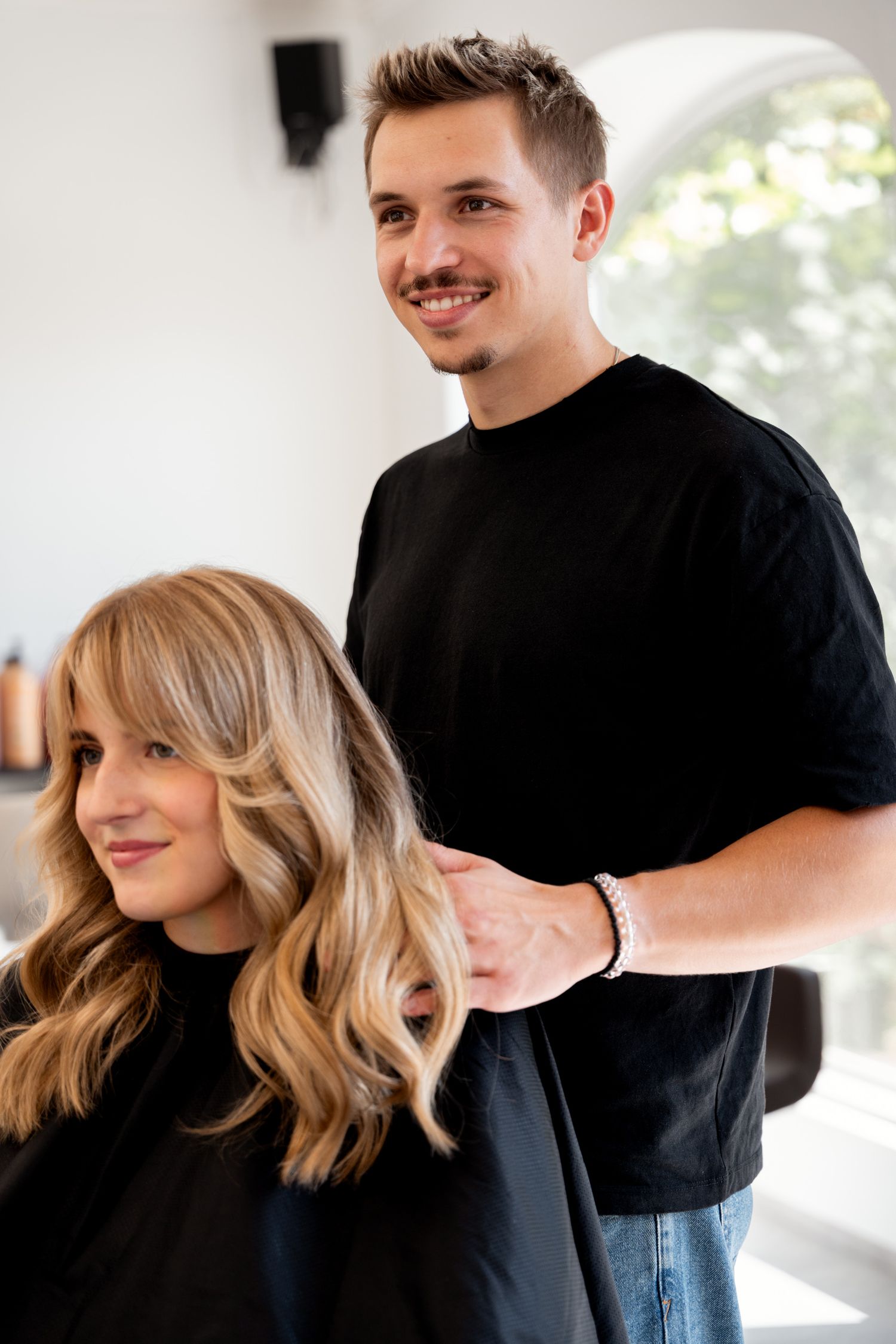 Male hairstylist in black shirt styling blonde client hair in bright modern salon.
