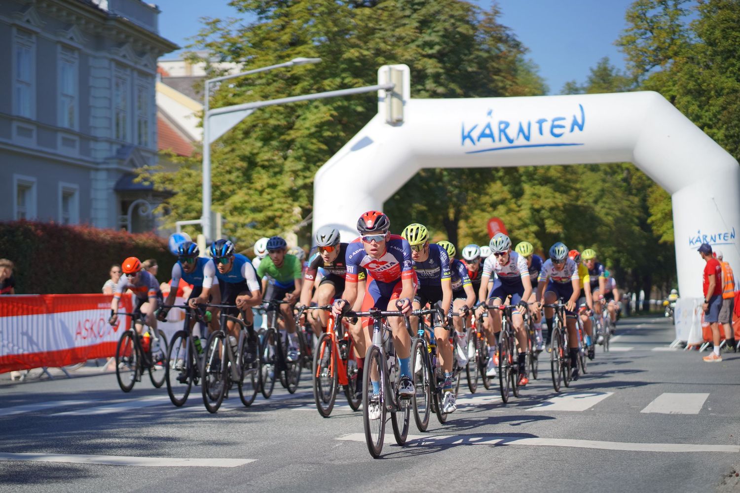 Cyclists racing under a Karnten inflatable arch at a sunny outdoor road cycling event.
