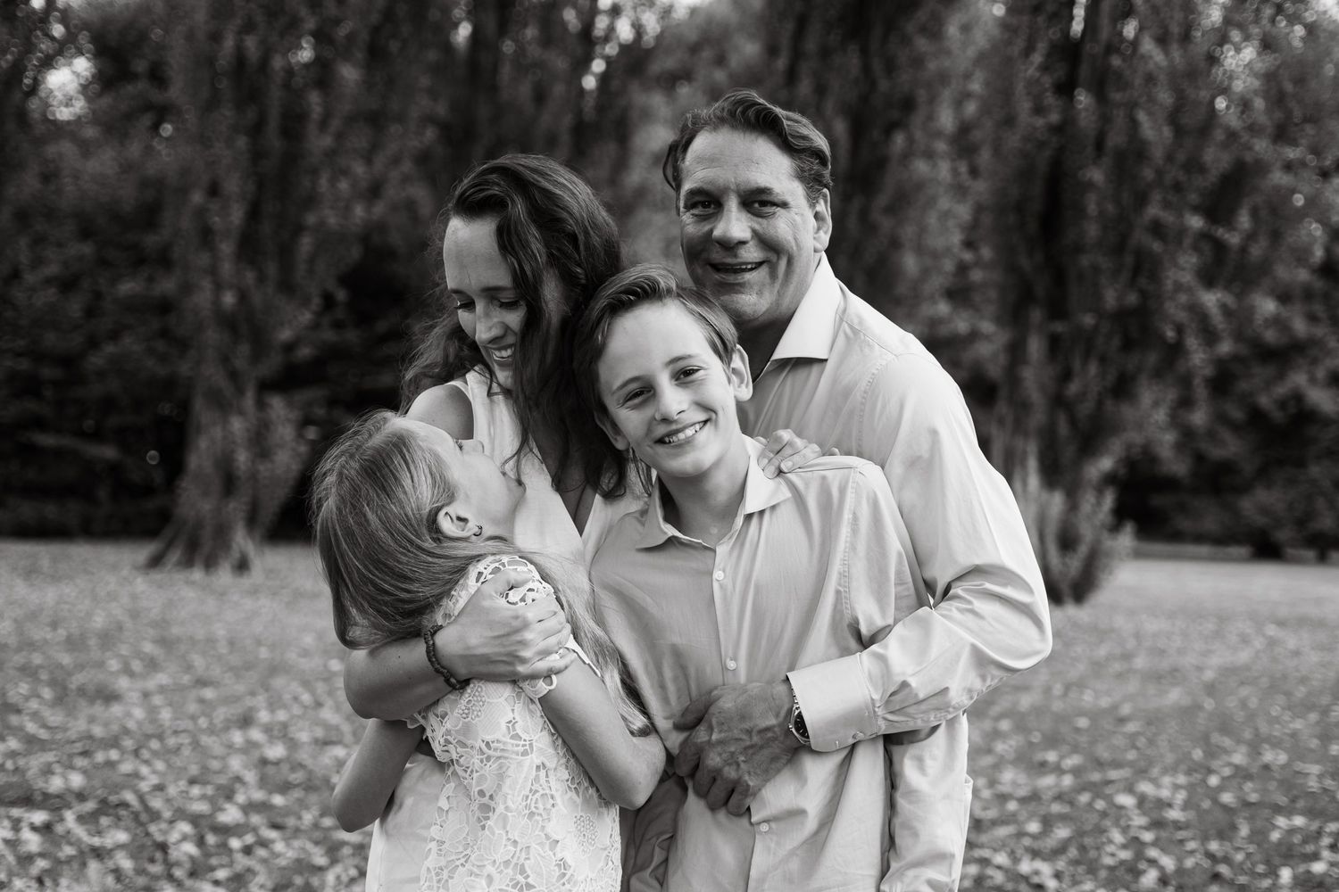 Black and white portrait of a joyful family of four hugging in a meadow.