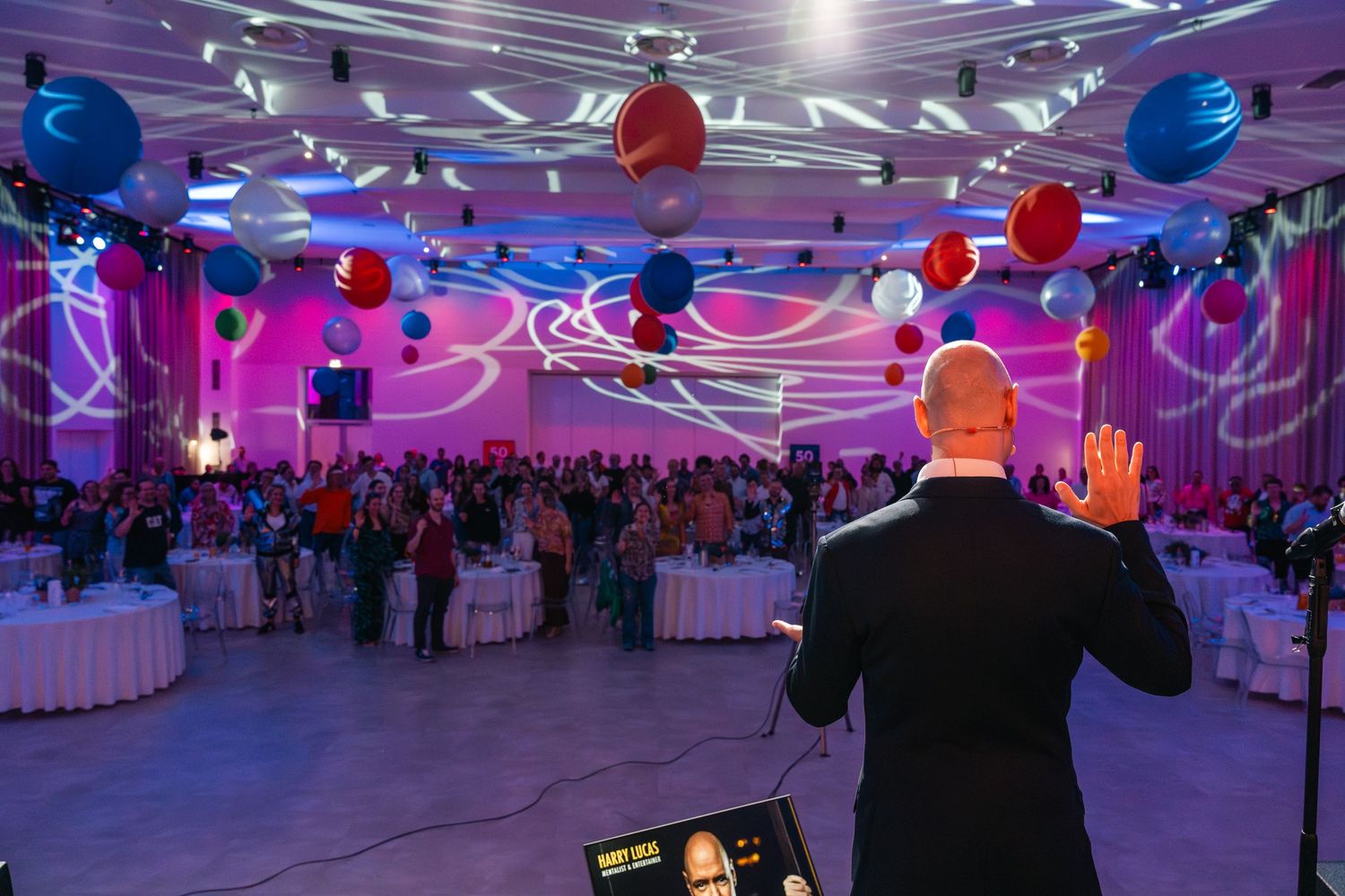 Speaker addresses a large crowd at a colorful corporate event with balloons and vibrant lighting projections.