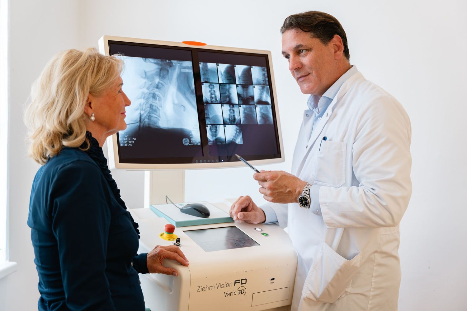 Male doctor in white coat discussing spine X-rays with female patient at medical imaging workstation.