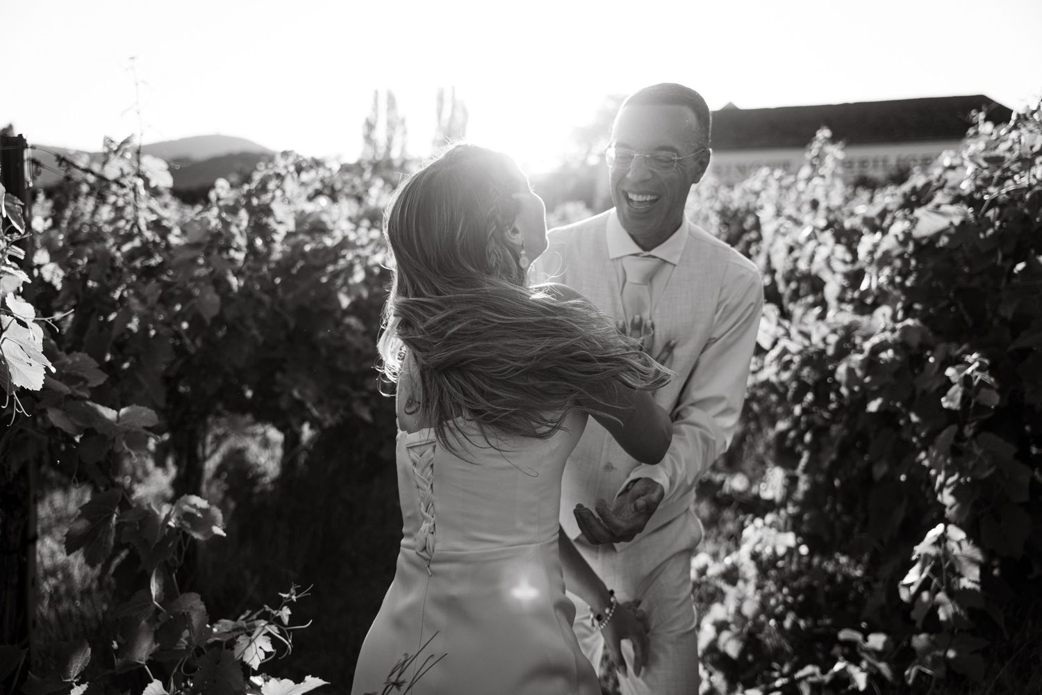 Black and white photo of bride and groom dancing joyfully in a sunlit vineyard.