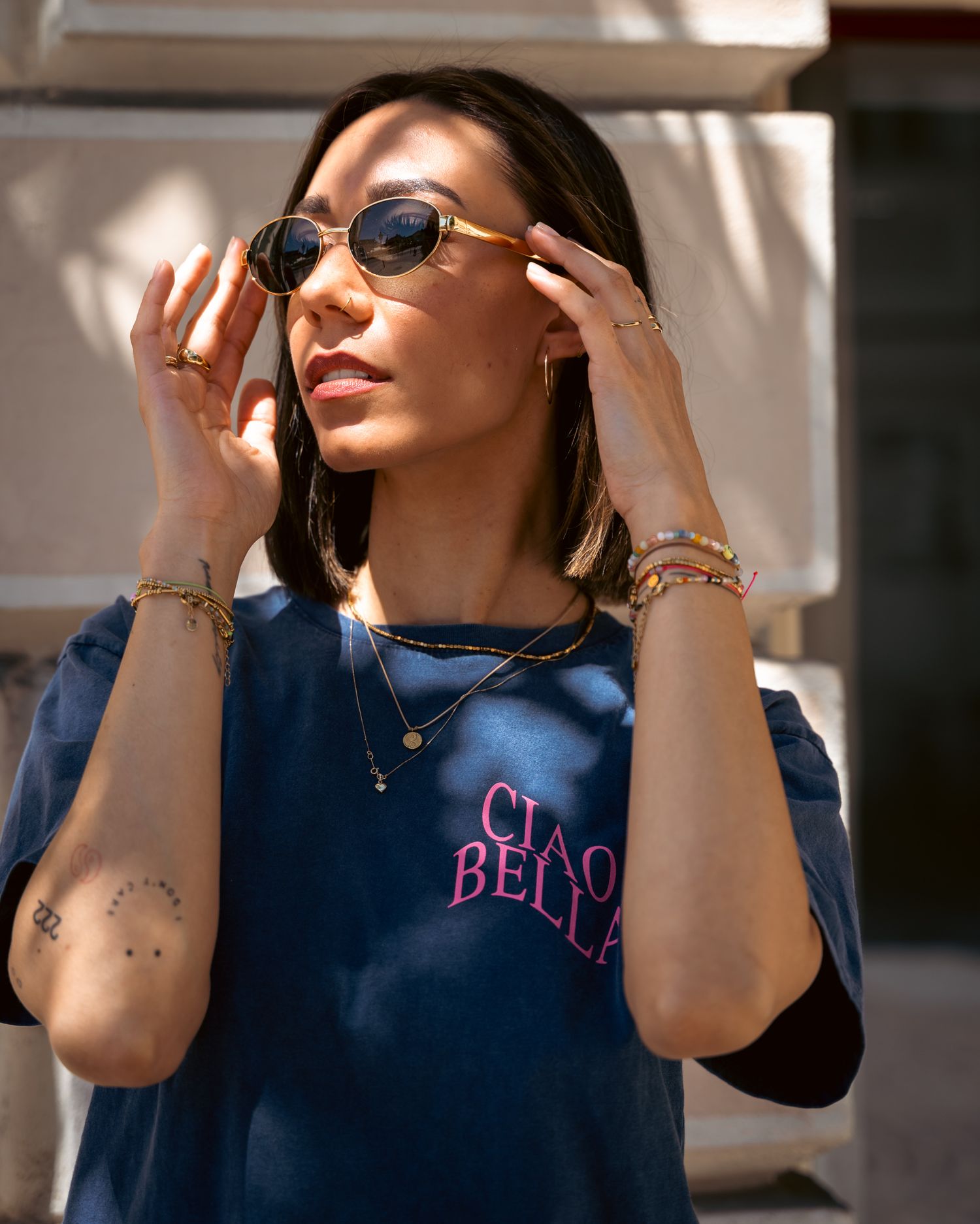 Stylish woman adjusting gold sunglasses wearing a Ciao Bella navy tee with layered necklaces.