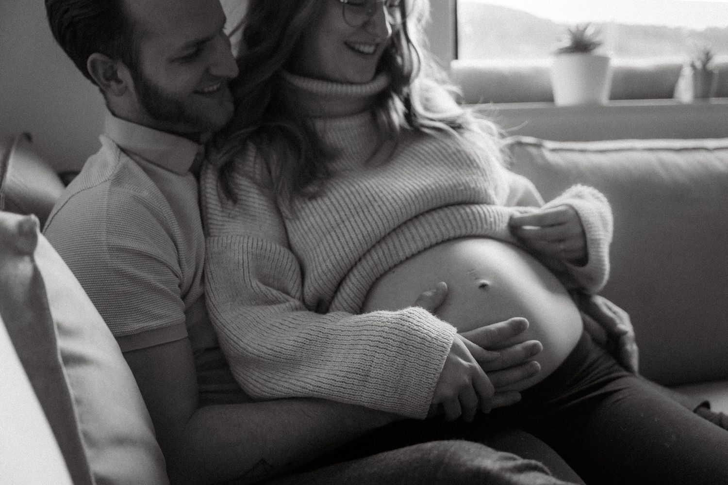 Couple sitting on a couch, man embracing pregnant woman with heart drawn on her baby bump, black and white photo.