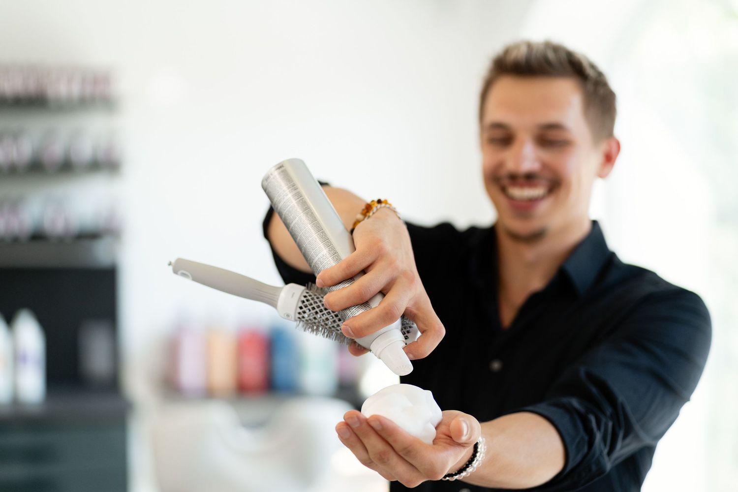 Smiling male hairstylist dispensing hair product from a pump bottle in a salon.