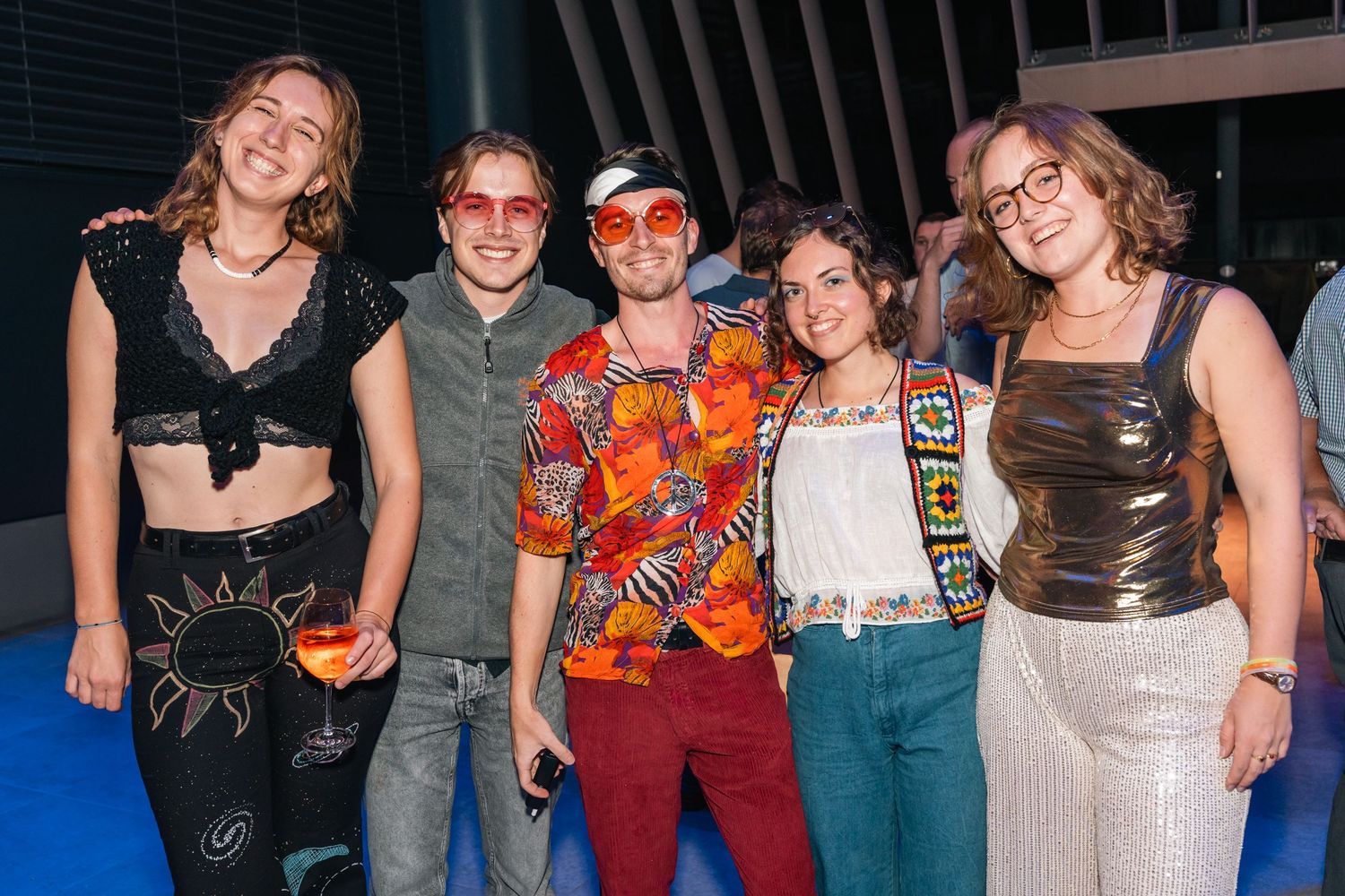 Group of five young friends posing together at a colorful outdoor evening social event.