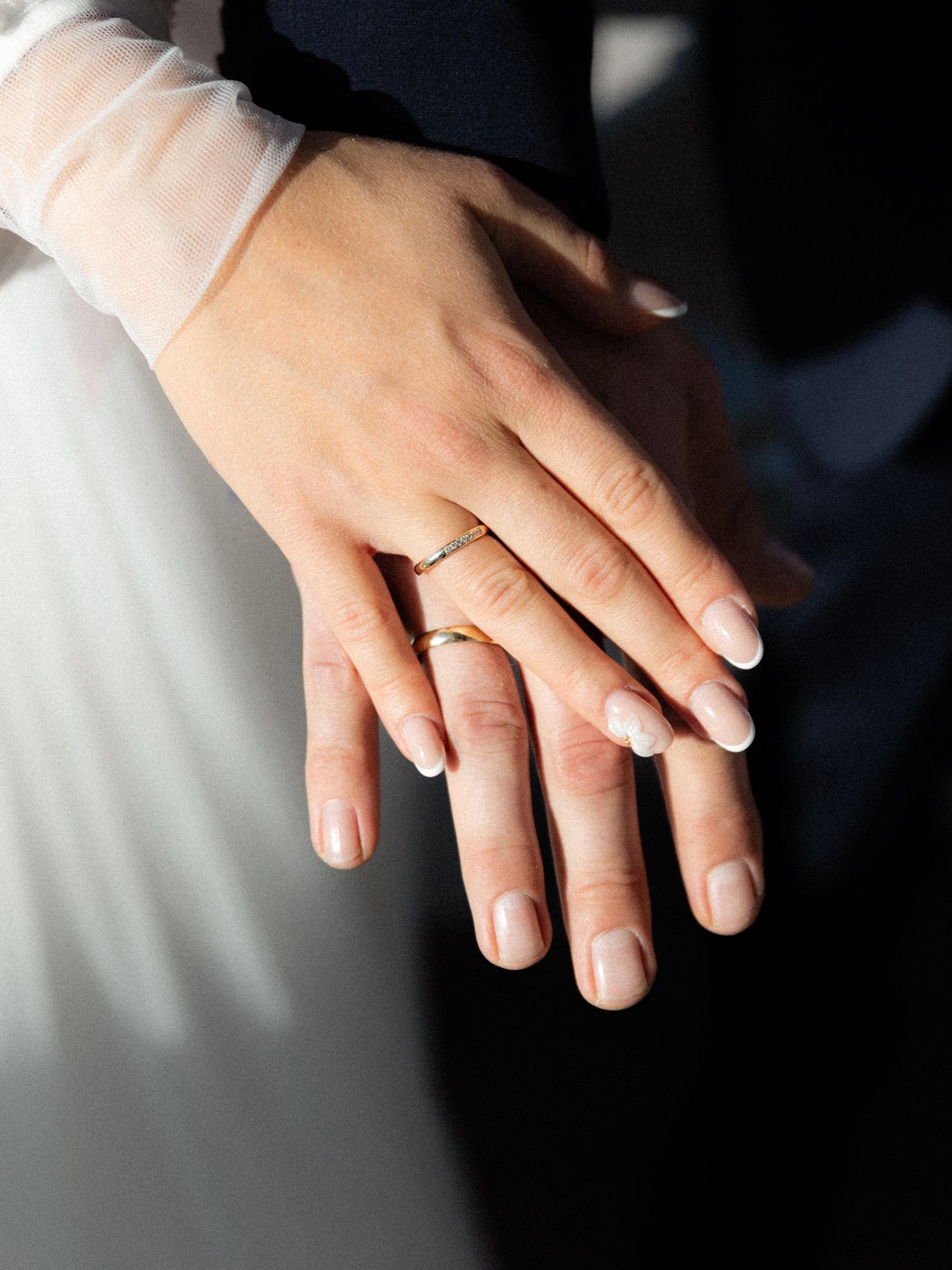 Close-up of newlyweds hands overlapping showing matching gold wedding bands against white dress fabric.