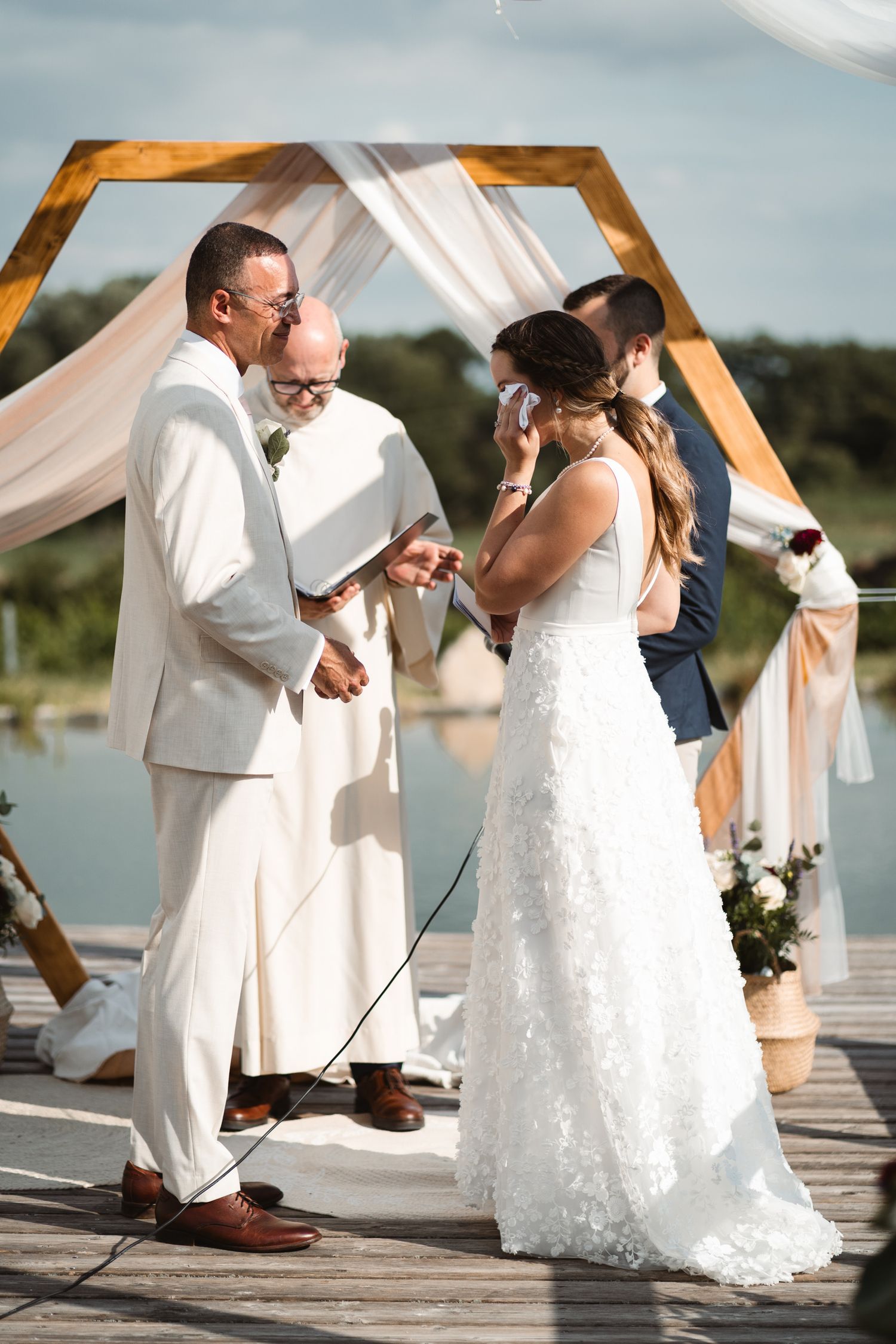 Emotional bride wiping tears during outdoor waterfront wedding ceremony under hexagonal arch.