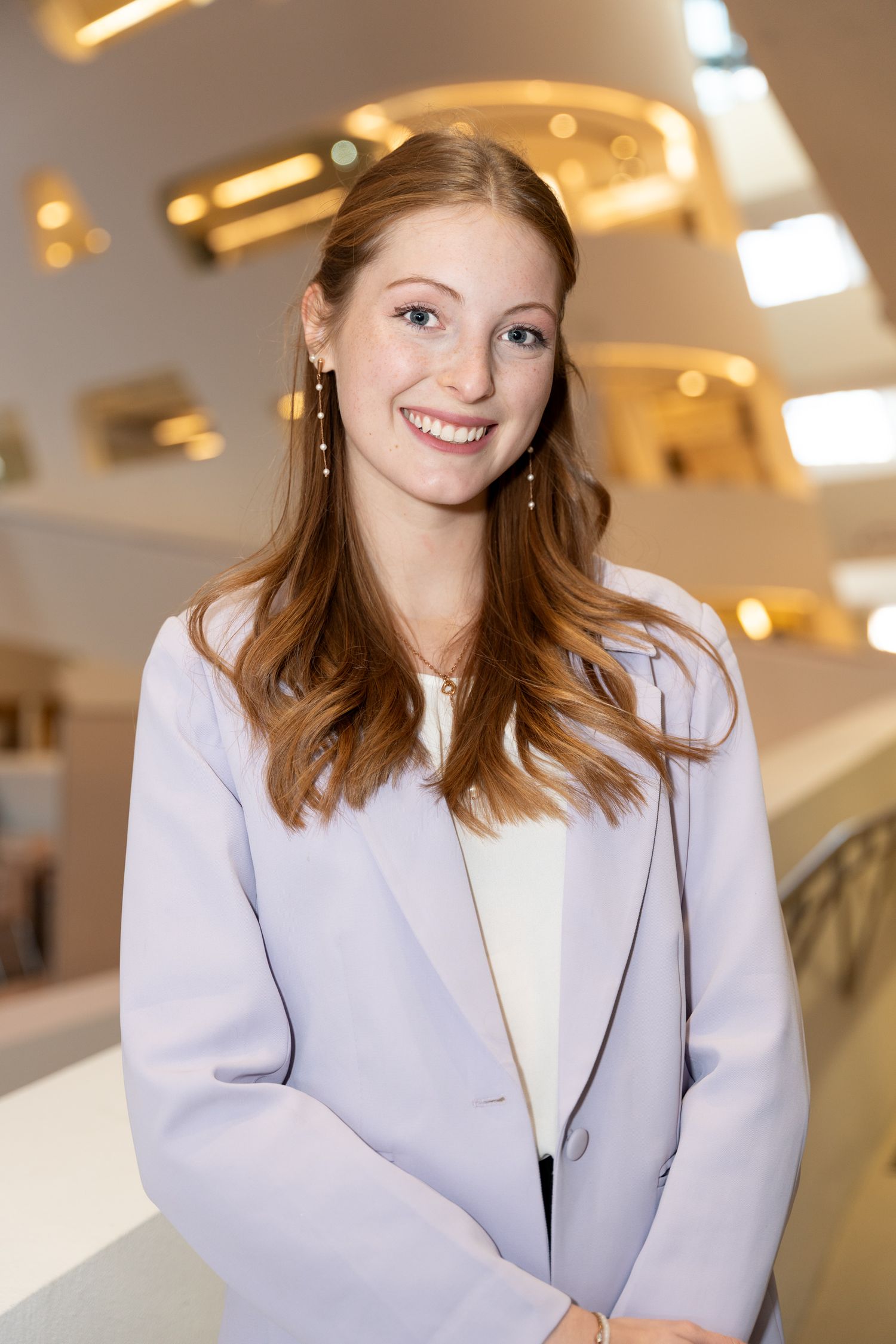 Smiling redhead woman in lavender blazer posing inside modern building interior.