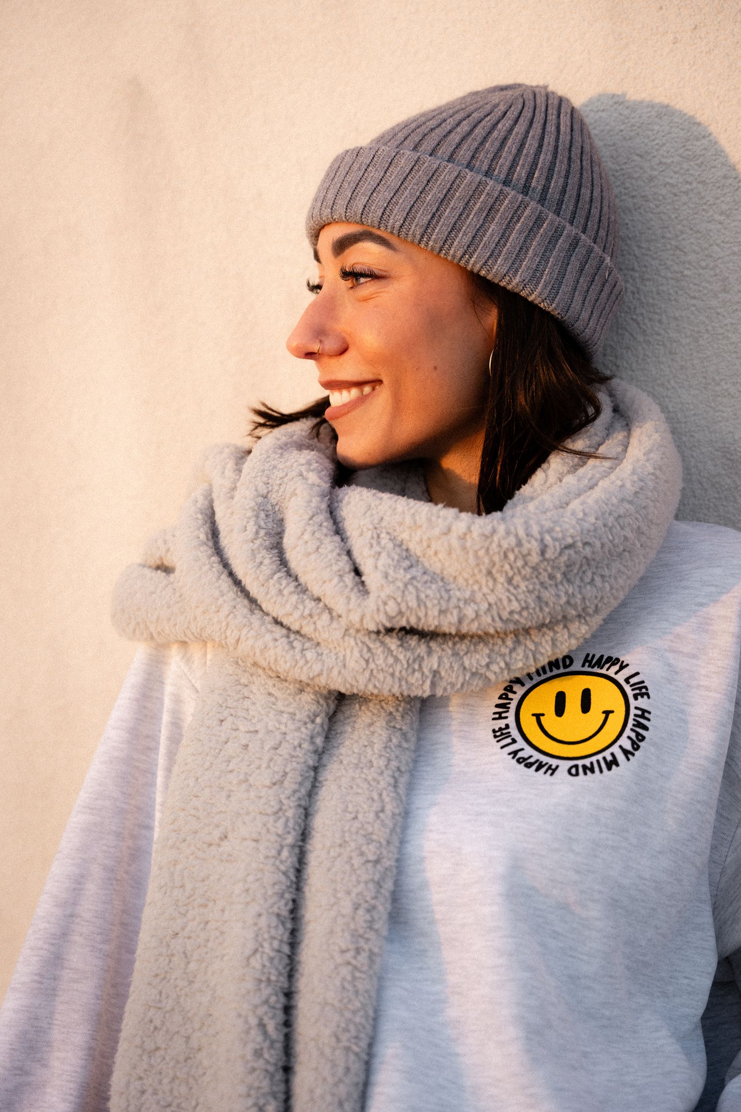 Happy young woman in grey beanie and fluffy scarf wearing smiley face hoodie.