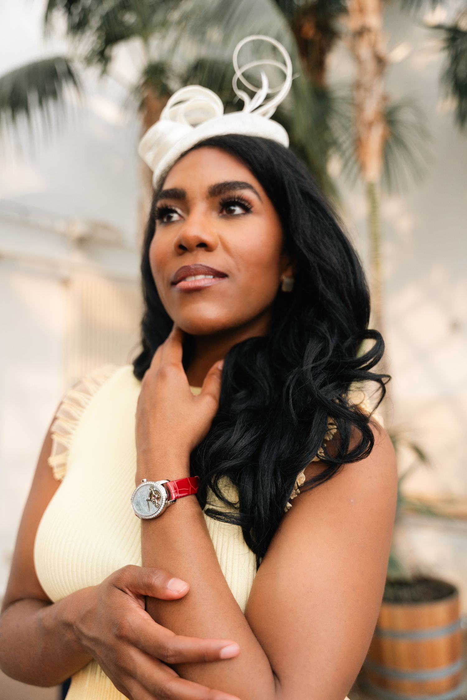Elegant Black woman in yellow dress wearing luxury watch and white fascinator hat.
