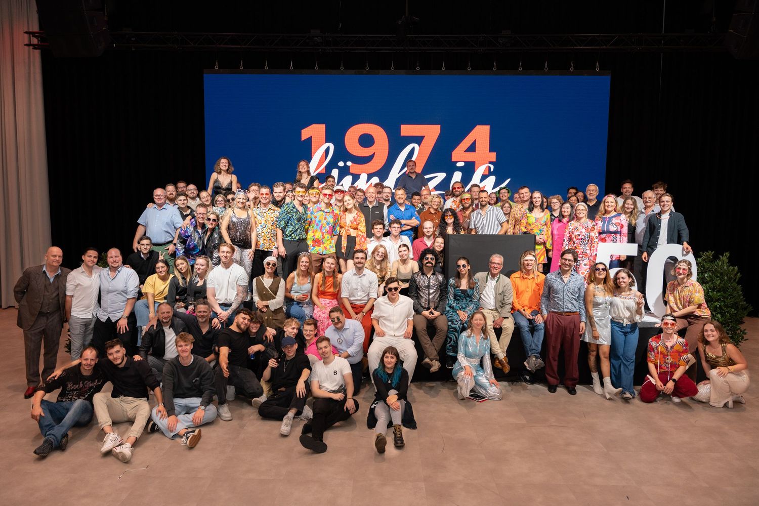 Large group photo of people posing on stage in front of a colorful 1974 anniversary backdrop.