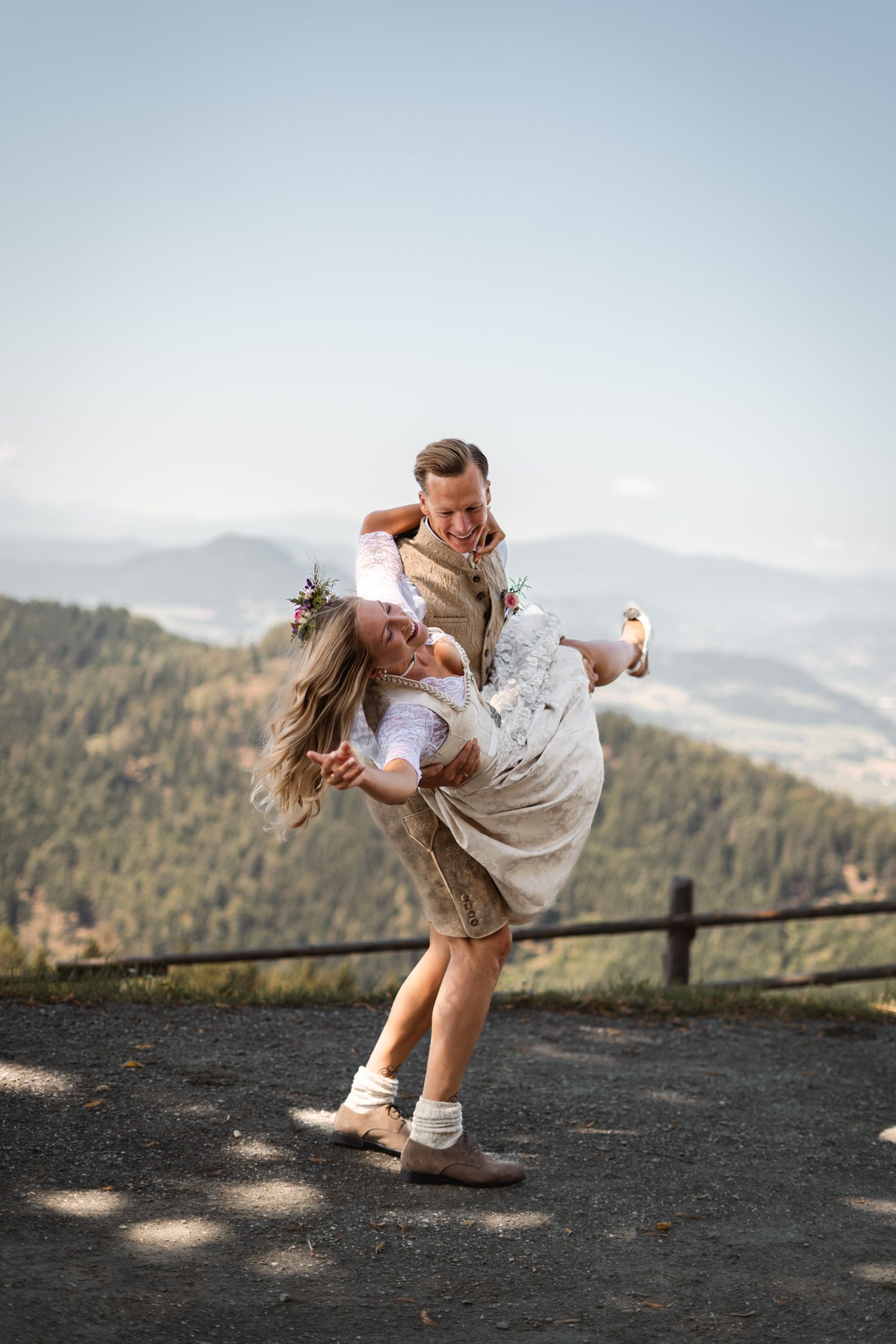 Joyful groom lifting bride in lace dress on mountain viewpoint with scenic valley backdrop.