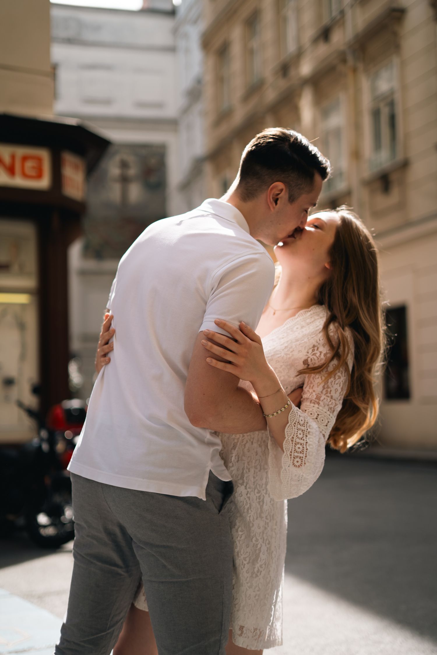 Romantic couple kissing on a sunlit European city street, dressed in casual summer outfits.