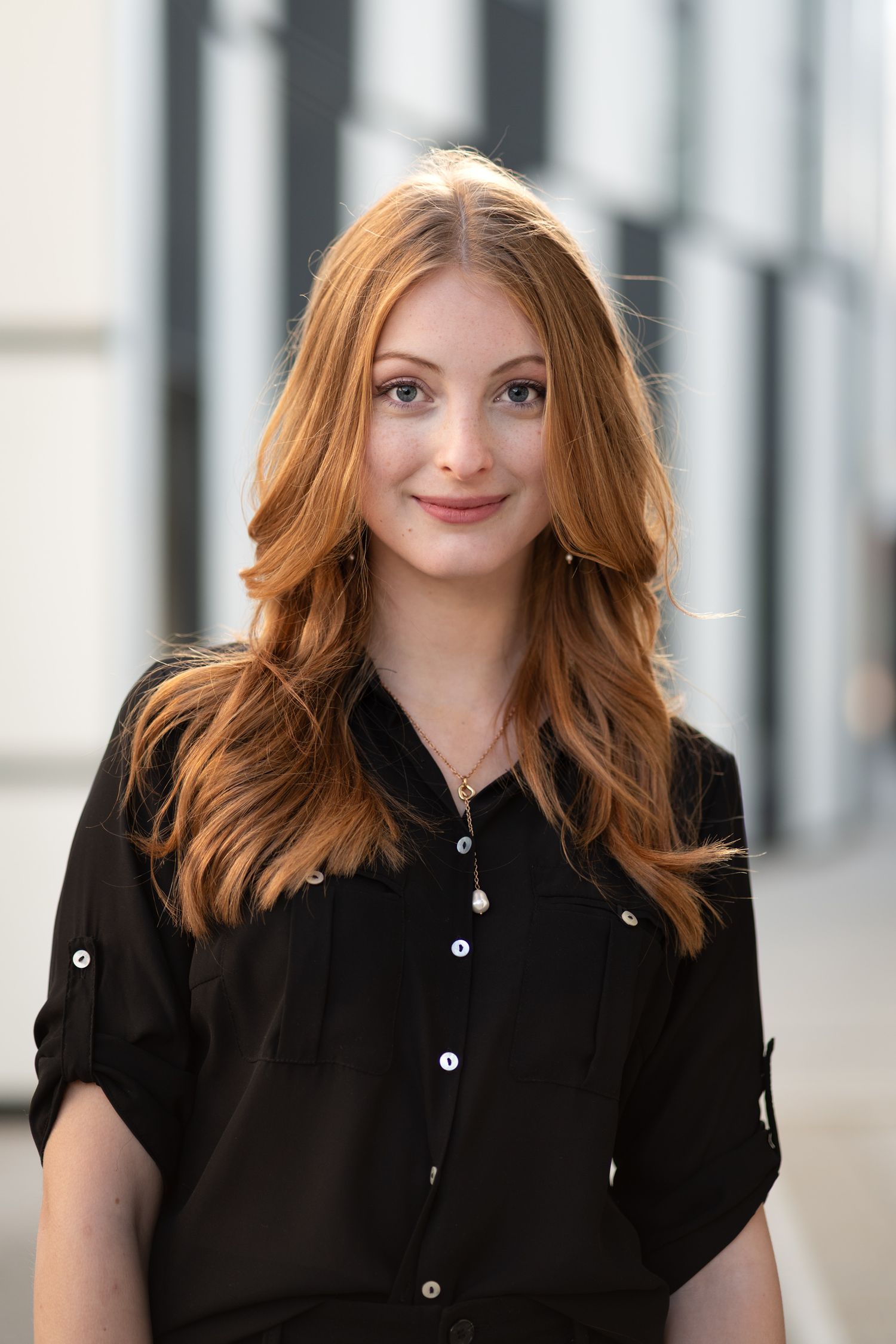 Young woman with long red hair smiling outdoors wearing black shirt in urban setting.
