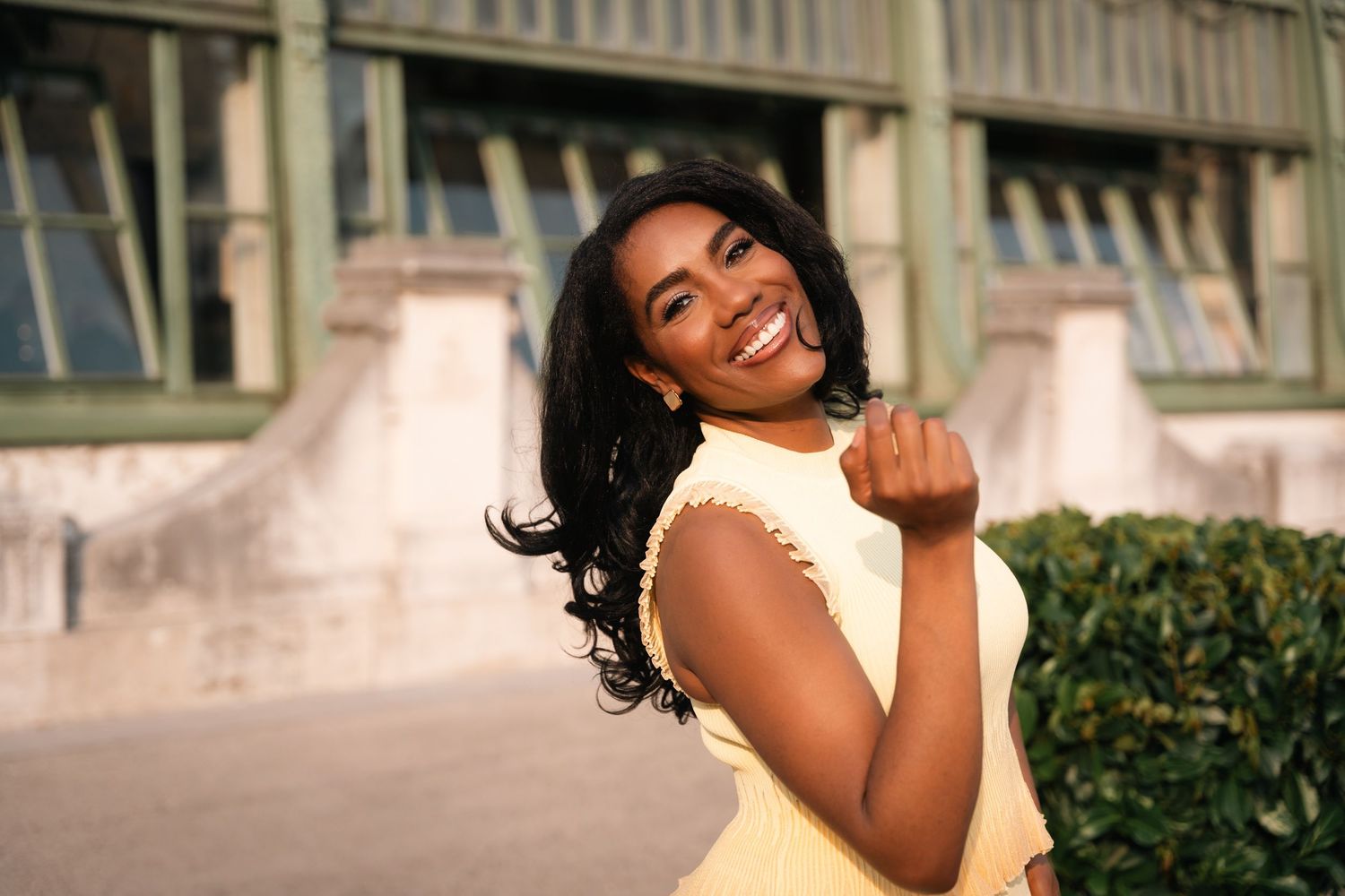 Smiling young Black woman in a yellow dress posing outdoors near green hedges and a building.