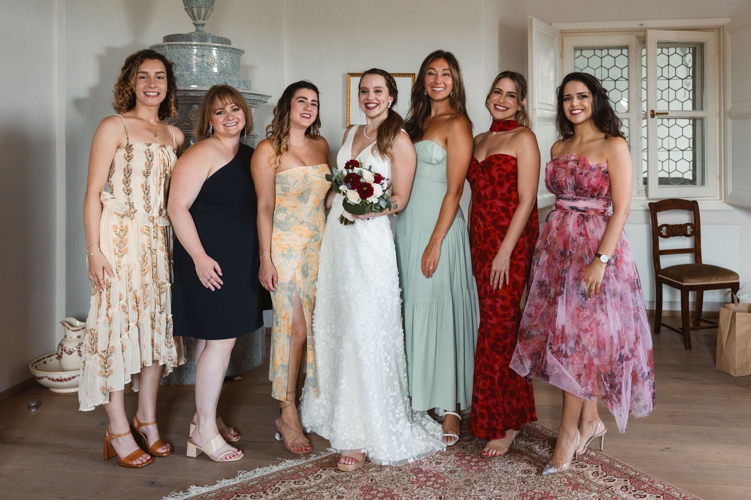 Bride posing with six bridesmaids in colorful dresses holding a burgundy floral bouquet indoors.