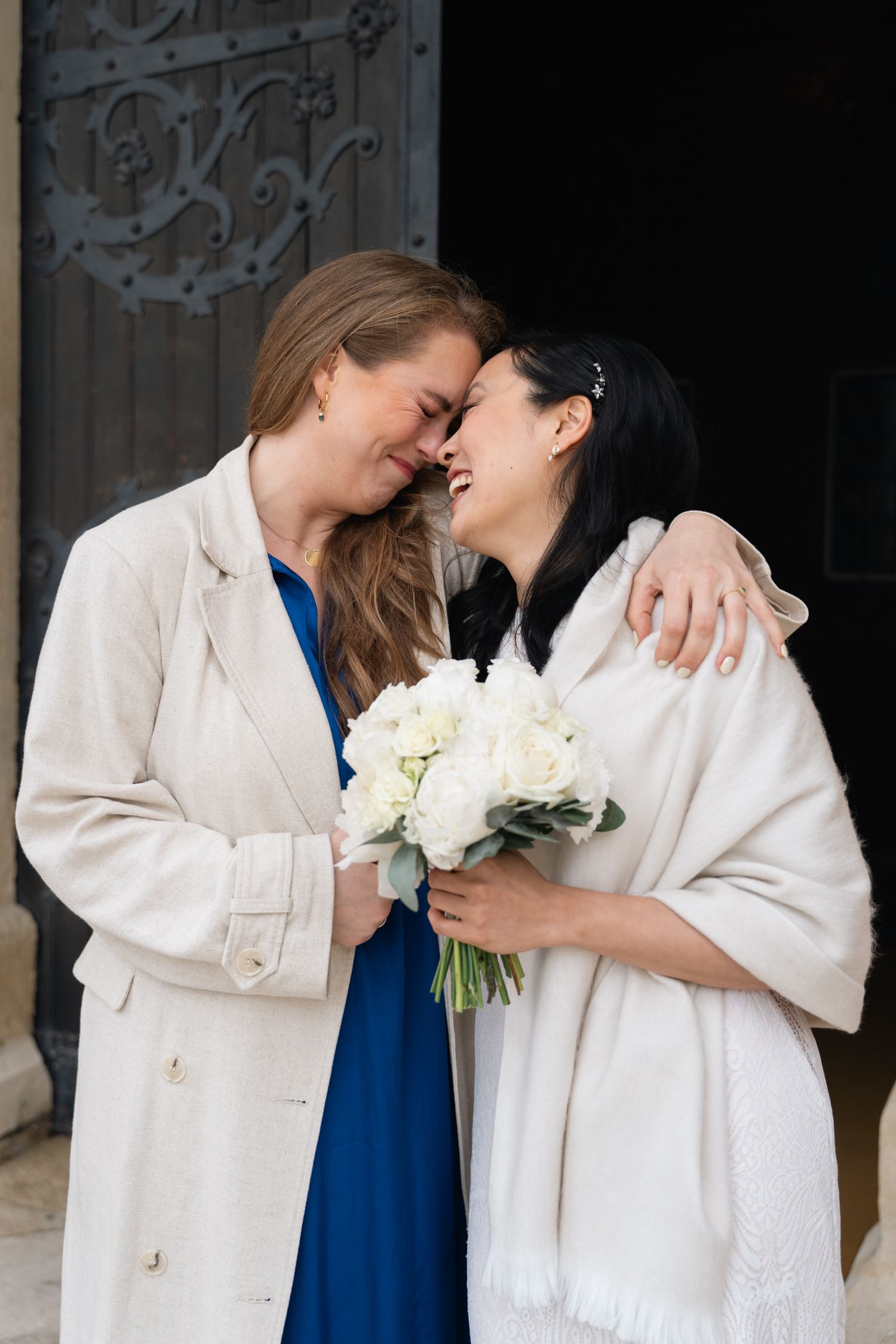 Two brides share a tender forehead touch holding white roses bouquet at church door.