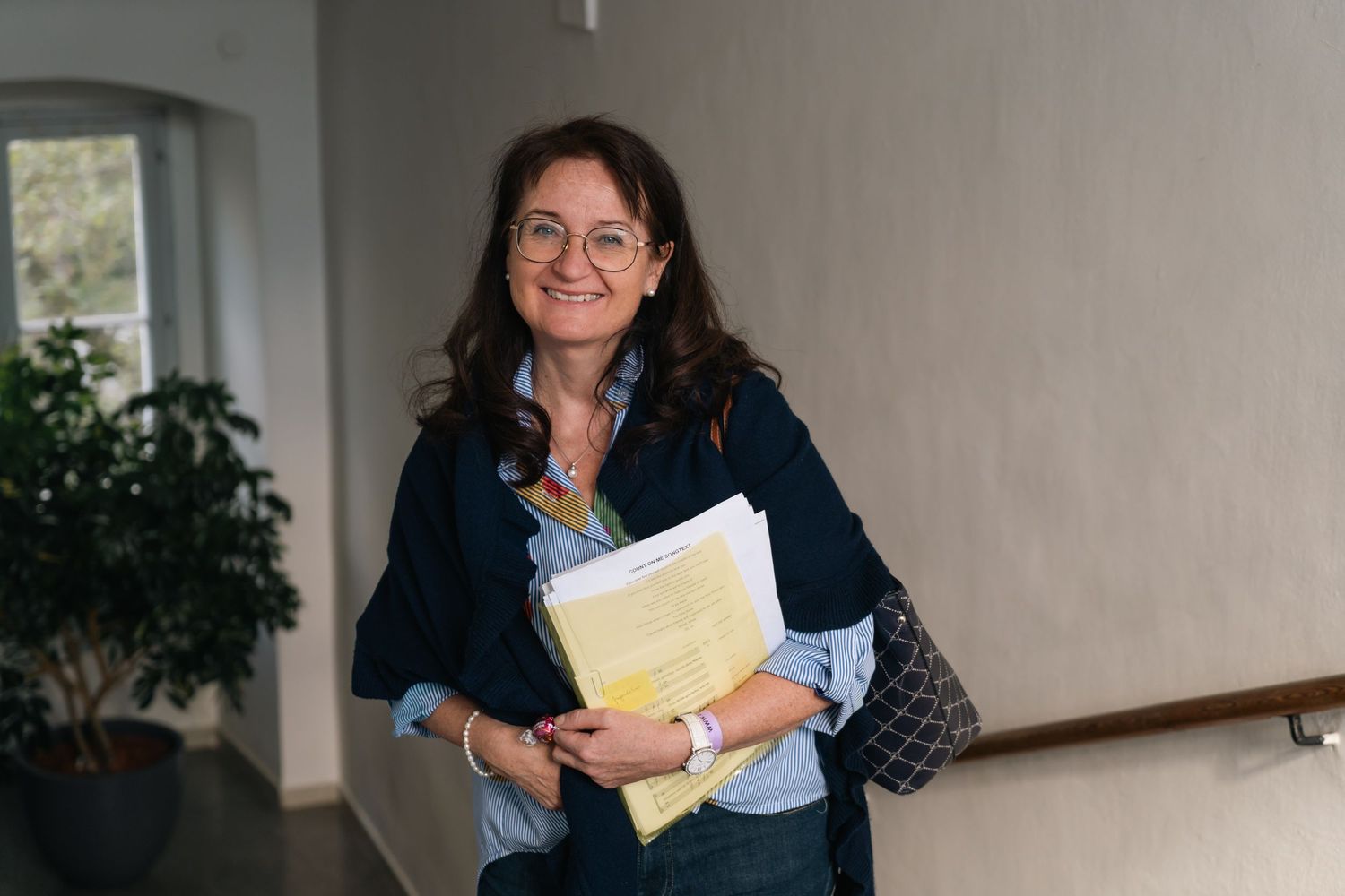 Smiling woman with dark hair holding yellow documents while standing on a staircase indoors.