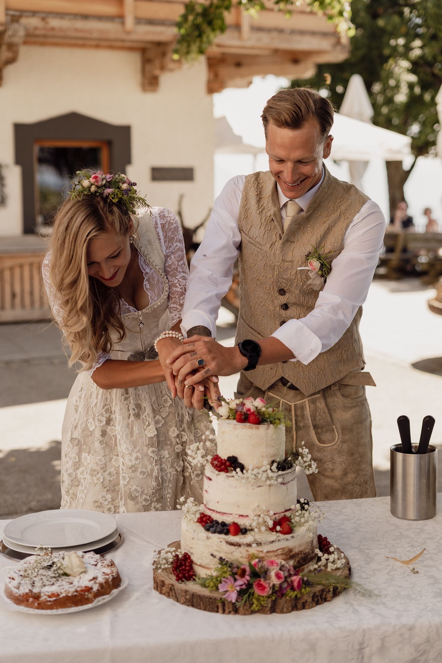 Bride and groom cut three-tier floral wedding cake outdoors together.