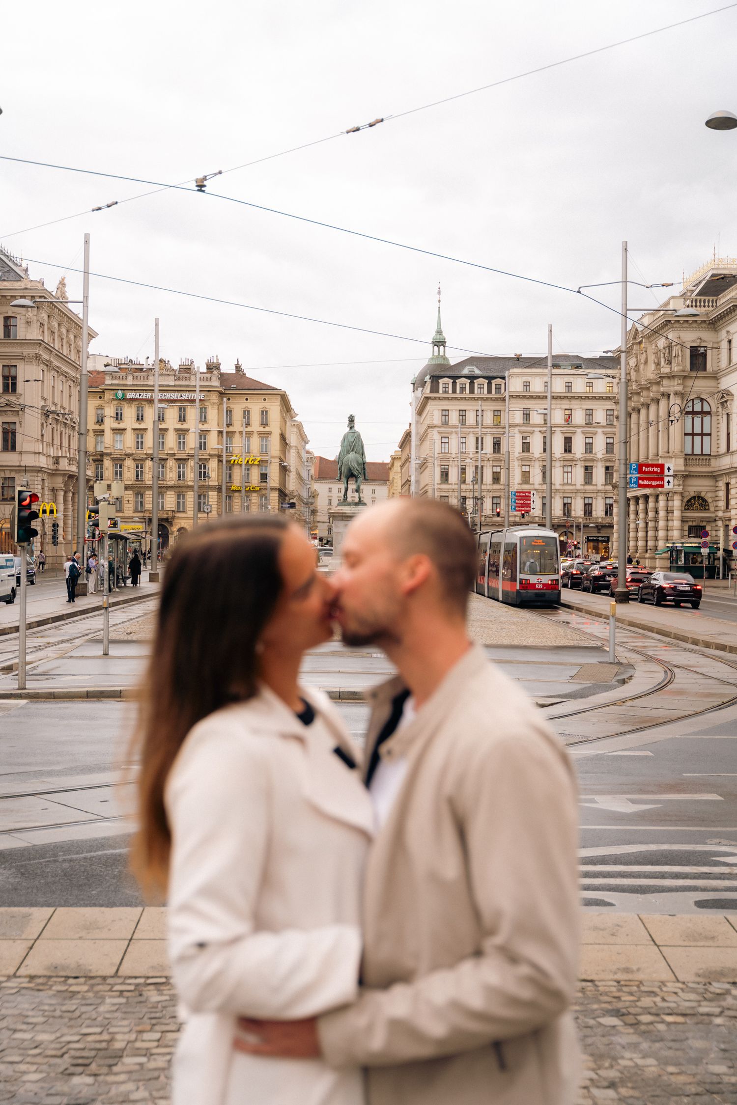 Couple sharing a kiss in a blurred foreground with Vienna city square and statue behind them.