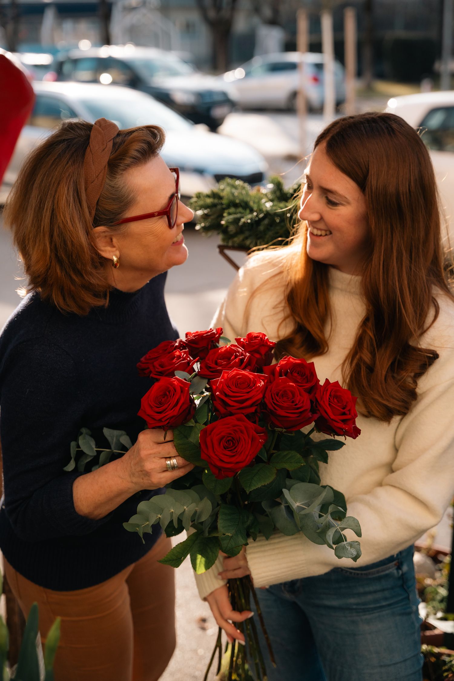 Two women smiling and sharing a bouquet of red roses with eucalyptus outdoors on a sunny day.