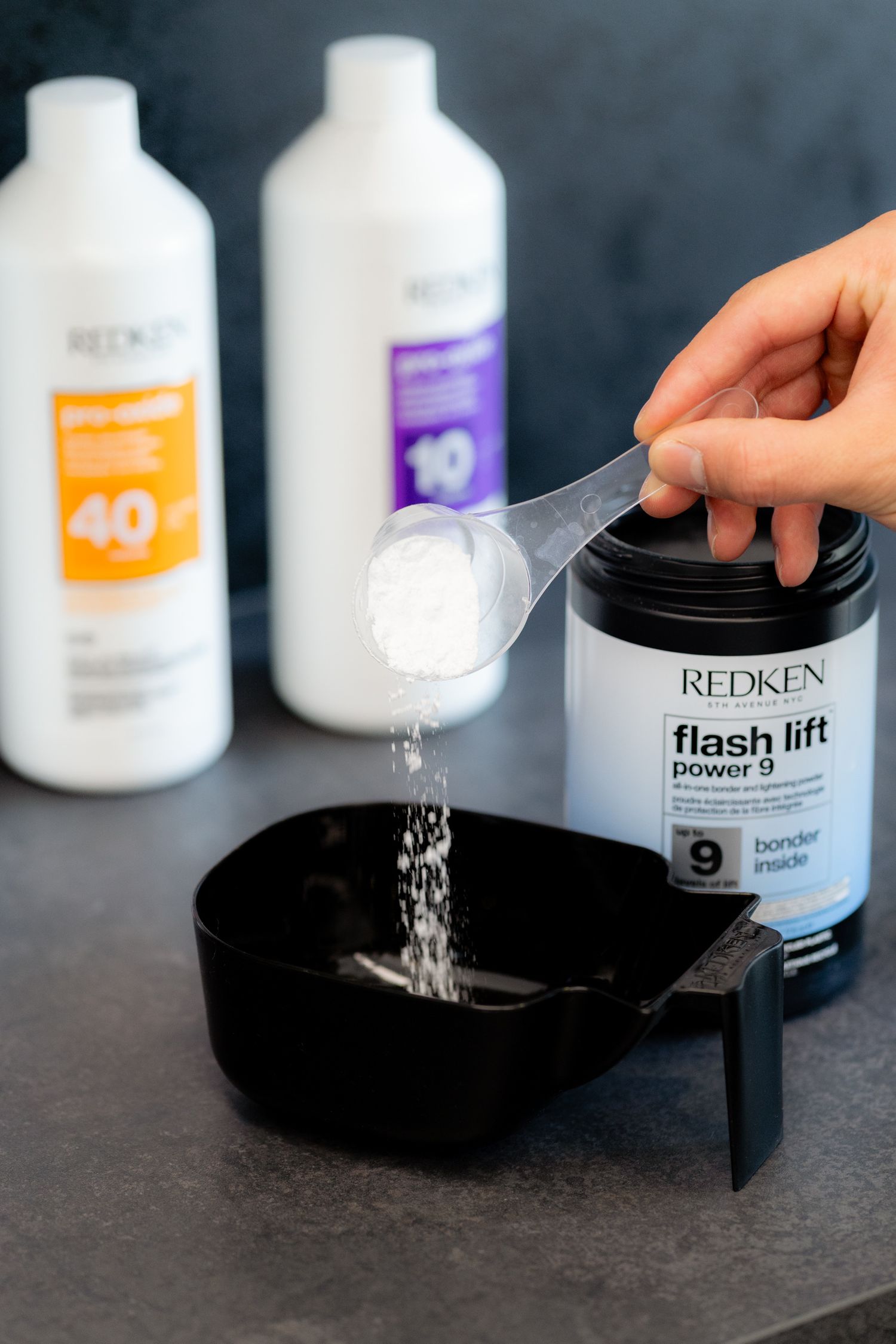Redken Flash Lift Power 9 bleach powder being measured into black mixing bowl with developer bottles.