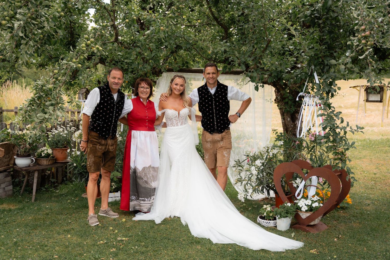 Bride in white gown posing with four family members under an apple tree at outdoor wedding.