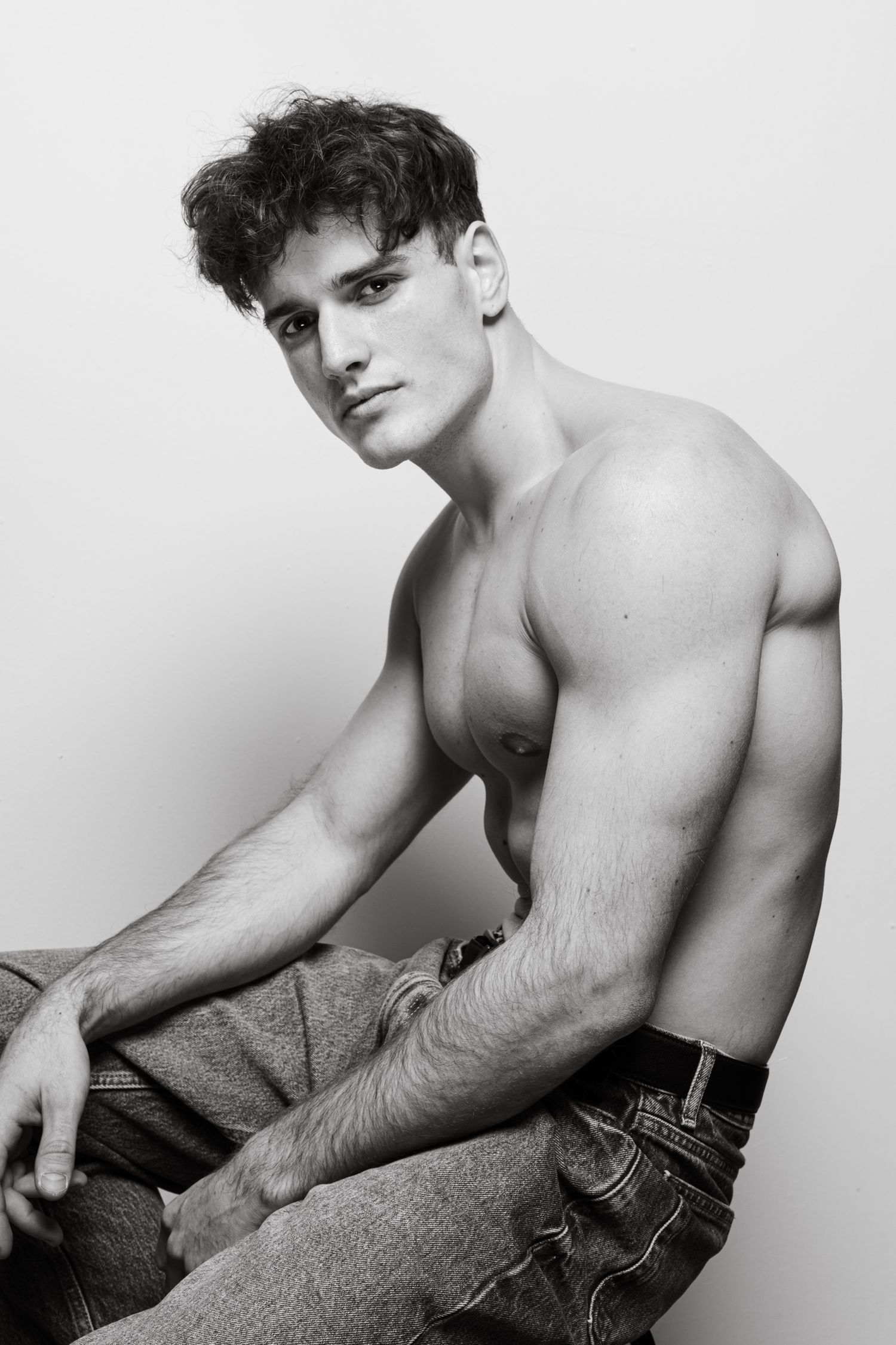 Black and white photo of muscular shirtless man in jeans sitting casually.