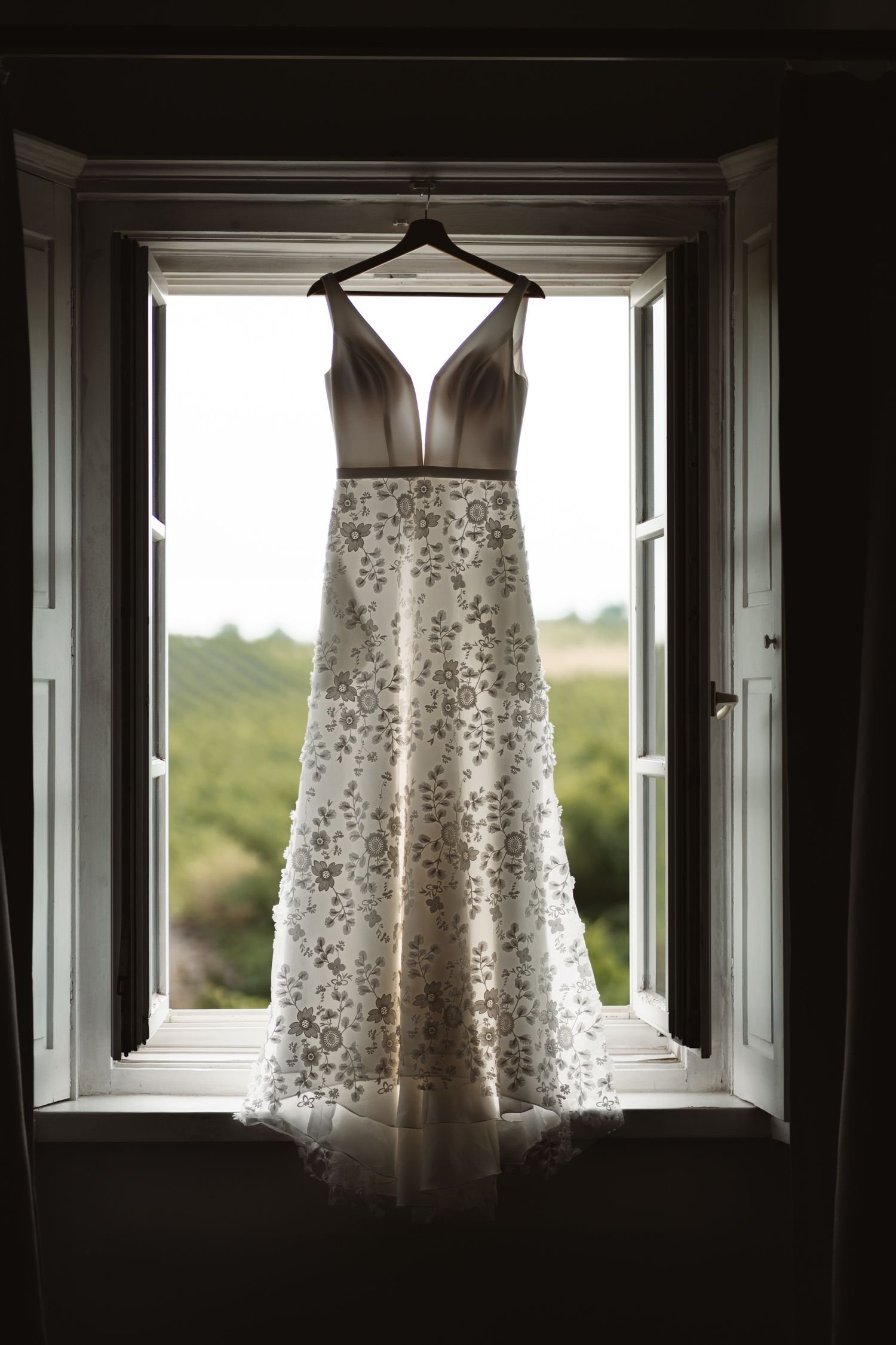 Elegant floral wedding gown hanging on a wooden hanger in an open window with a green countryside view.