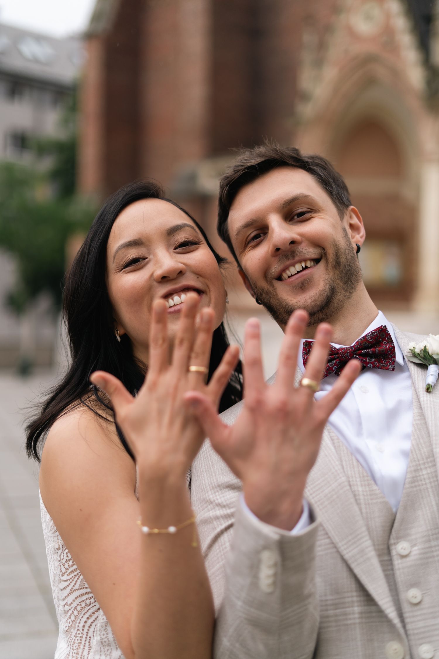 Smiling newlyweds showing off wedding rings to camera in front of a historic church.
