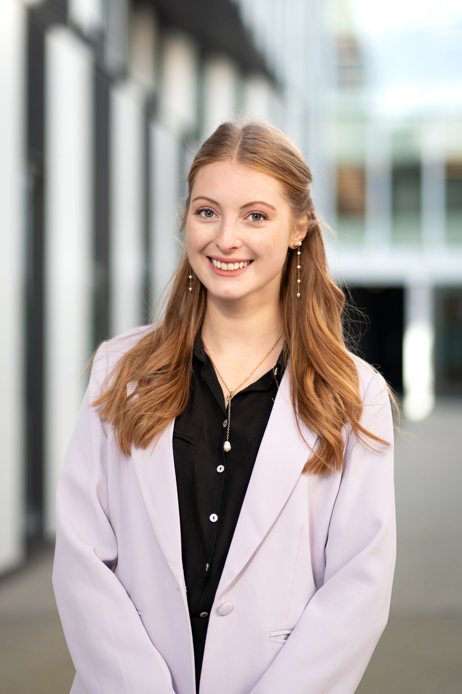 Cheerful young woman with long auburn hair wearing a lavender blazer, smiling outdoors near a building.