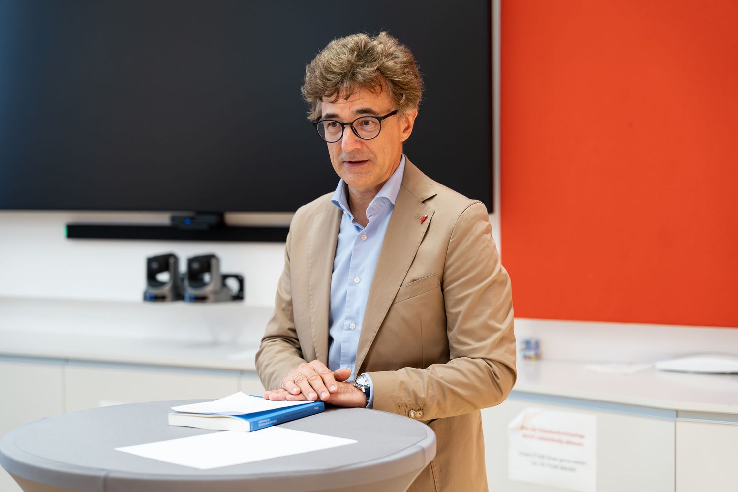 Man in beige blazer speaking at a round white podium in a modern conference room with orange wall.