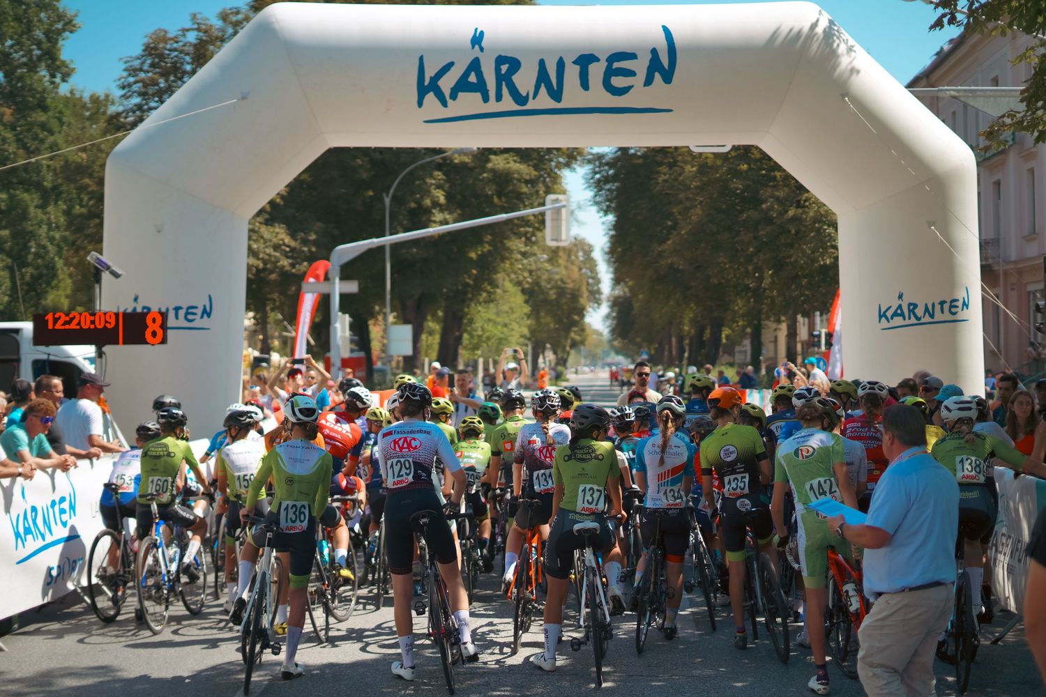 Cyclists lined up at a Kärnten branded inflatable start arch during an outdoor road cycling race.